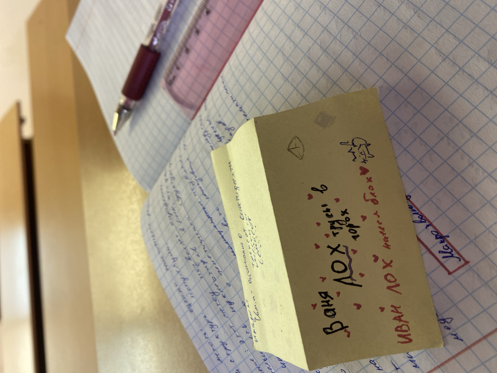 A close-up shot of a page from a notebook, with handwritten notes and a small, folded piece of paper on top. The notebook page contains mathematical equations and formulas written in Cyrillic script. The folded paper is yellow and also has handwritten Cyrillic text and small heart drawings on it. The text on the paper appears to be a message or note. In the background, a pen and ruler are visible, adding to the context of the scene. The overall environment suggests a learning or study setting.