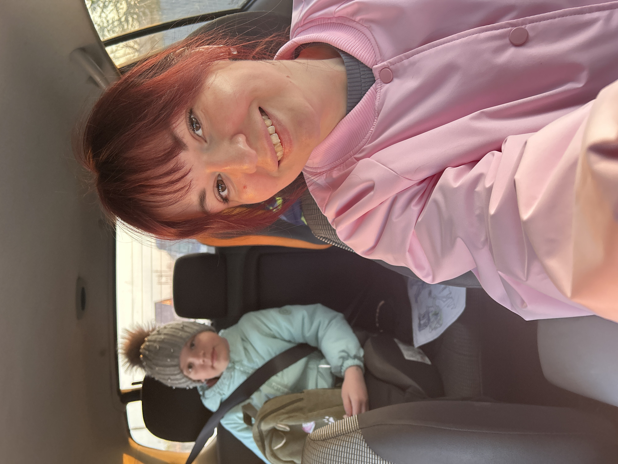 The image shows a woman and a child in the back seat of a vehicle, possibly a car. The woman is facing the camera, smiling, and wearing a pink jacket. The child, wearing a winter hat and a light blue jacket, is also facing the camera but appears more relaxed. Both are wearing seatbelts. The interior of the car is visible, including the seats and a window with a blurred background suggesting the vehicle is in motion. The location Фоки, Russia is not visible in the image.