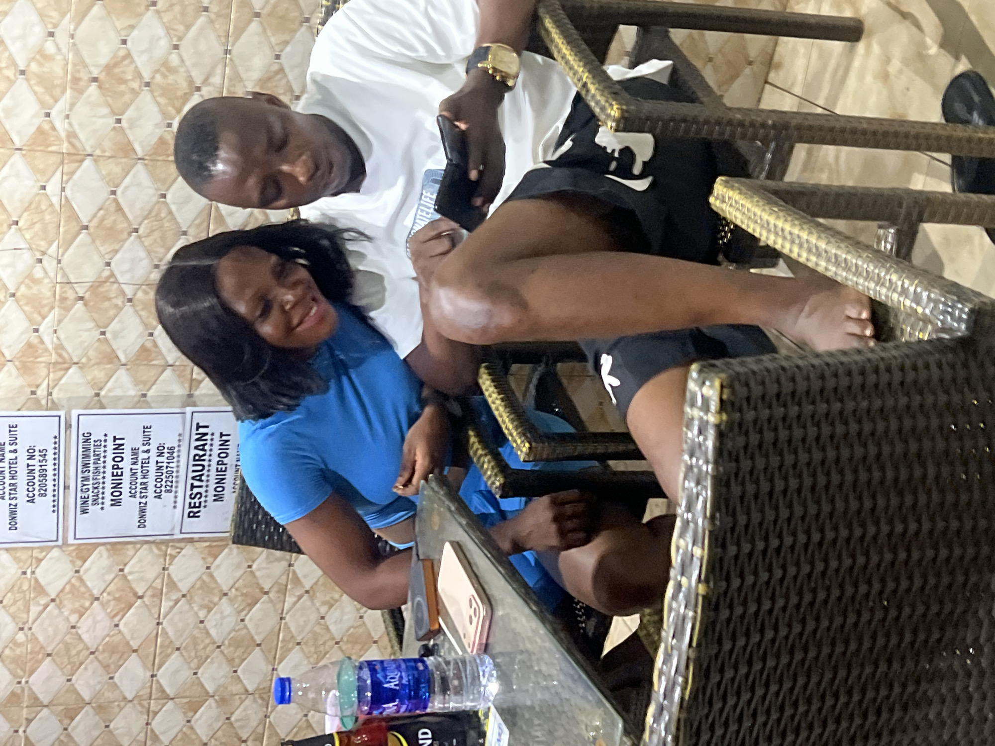 Here's a forensic description of the image:
**Scene:** The photo appears to be taken indoors at the "Restaurant Moniepoint" located in Nkpor, Nigeria, part of the "Donwiz Star Hotel & Suite" complex. The interior features tiled walls with signs displaying account details for various services, including dining, wine, gym, swimming, and snacks.
**People:** A man and a woman are seated at a table. The man is wearing a white shirt, black shorts with a white design, and a wristwatch. He is looking at his phone. The woman is wearing a blue top and short. She is looking towards the camera and smiling.
**Objects:** On the table are at least two phones, a bottle of water, and another bottle. The table and chairs appear to be made of woven material.
**Actions/Interactions:** The man is focused on his phone, while the woman is engaging with the camera. The relationship between them is unclear from the image alone.
**Environment:** The scene suggests a casual dining or socializing environment within the Moniepoint Restaurant at the Donwiz Star Hotel & Suite in Nkpor.