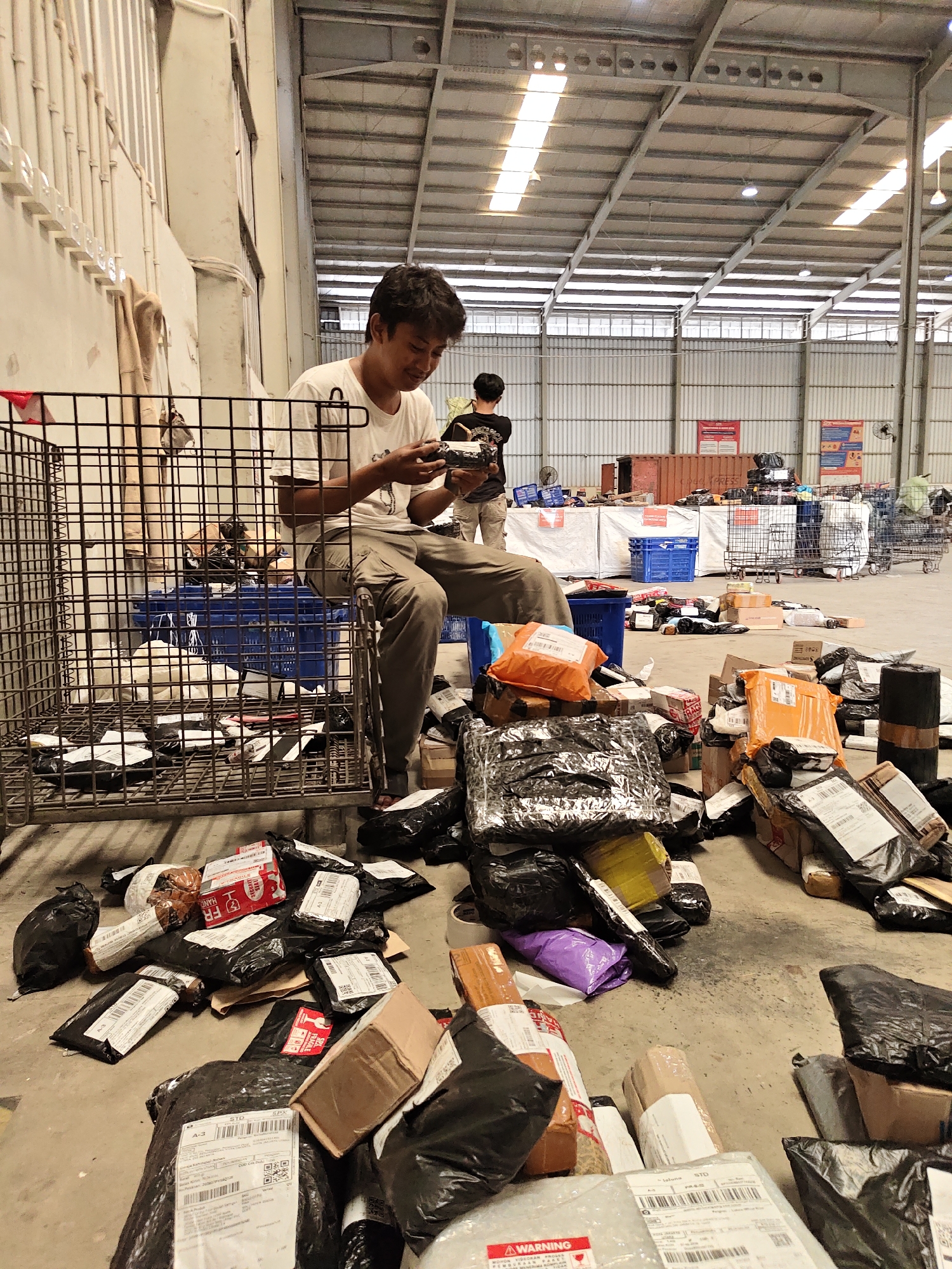 A young man in a white t-shirt and khaki pants is sitting on a metal cage in a warehouse in North Jakarta, Indonesia. He is holding a small electronic device and looking down at it. The warehouse is filled with packages of various sizes and shapes, most of which are black plastic bags. Some packages are inside the cage with the man, while others are scattered on the floor around him. In the background, there are other people, shelving units, and more packages. The overall impression is of a busy logistics or distribution center.