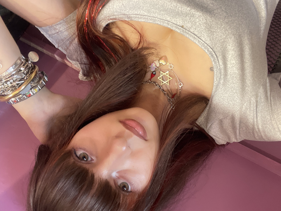 An upward-facing, close-up shot captures a person lying down or leaning back on a solid purple or magenta surface. The image is oriented with the person's head at the bottom center, gazing directly into the camera.
The person has long, dark brown hair with straight bangs covering their forehead. Notable features in their hair include streaks of shimmering red tinsel or strands, particularly visible towards the upper left of the image. Their eyes are wide and a light brown or hazel color, framed by defined eyeliner. Their nose is small, and their lips are slightly parted, adorned with a glossy, natural pink or nude lipstick.
They are wearing a light grey, possibly heathered, casual top or t-shirt. A layered necklace is prominent around their neck. The most visible pendant is a gold-toned Star of David. Accompanying it are smaller charms, including a red evil eye bead, a gold heart, and a small, iridescent purple heart or cluster. Various silver-toned chains complete the layered look of the necklace.
Their left arm (viewer's right) is visible, resting above their head. This arm is accessorized with a significant stack of bracelets. These include several silver and gold-toned bangles and chain bracelets. One distinct bracelet is a silver-toned charm bracelet featuring multiple colorful, rectangular inserts in shades of red, yellow, green, blue, and purple.
The background is a smooth, untextured, uniform purple or magenta color, suggesting an indoor setting on a flat surface. The lighting appears soft and even, highlighting the features of the person. The overall impression is a self-portrait or close-up candid shot, focusing on the individual and their accessories.