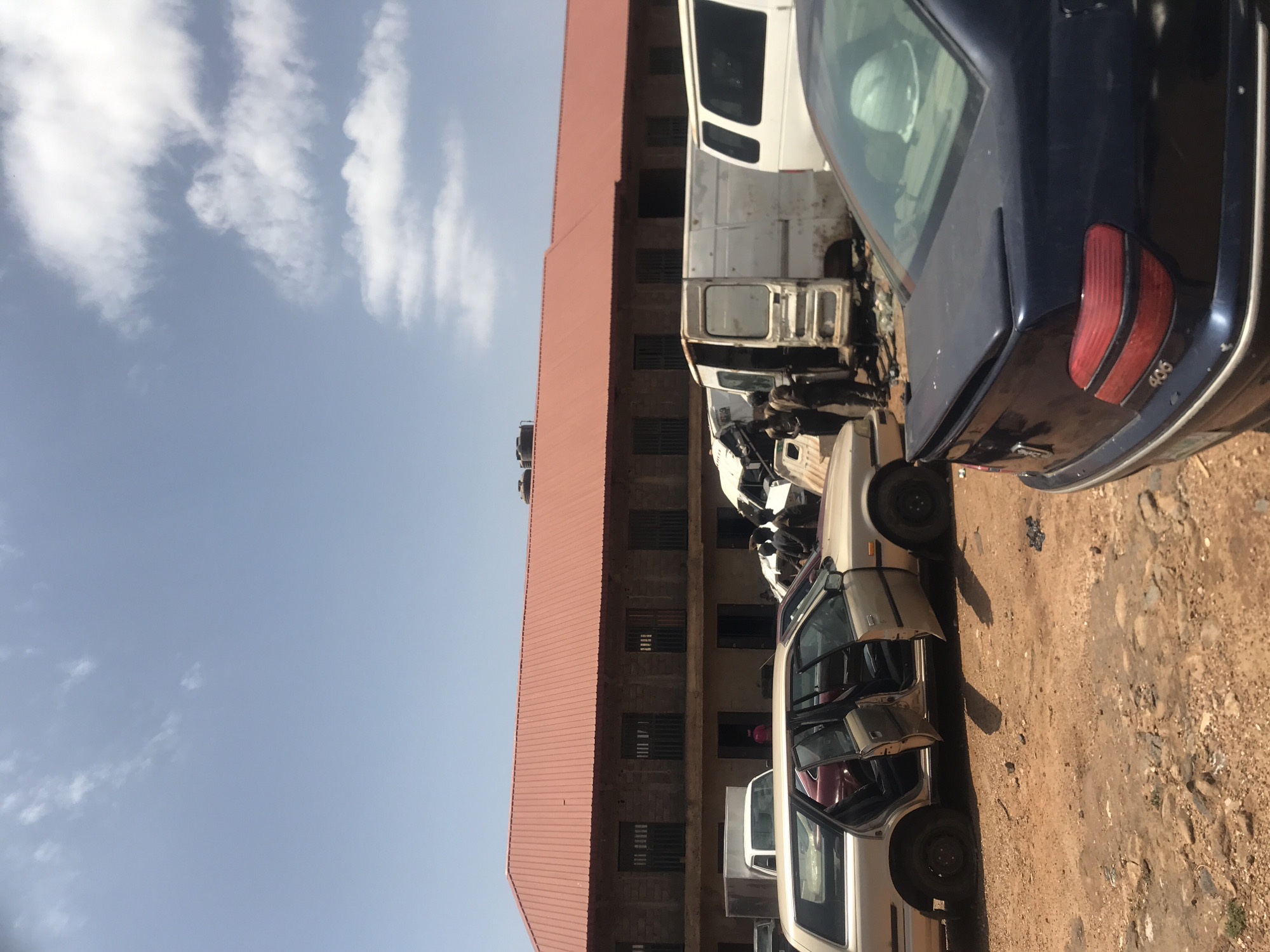 This image depicts an outdoor scene in Rafin Par, Nigeria, characterized by a large, two-story building with a red corrugated metal roof and barred windows. In front of the building, there is a collection of various vehicles, suggesting a car lot, repair shop, or junkyard. Among the vehicles are several vans, including white cargo vans, and multiple cars. One car, a tan station wagon with its doors open, is prominently featured in the foreground. Another car, a dark blue sedan with a visible "406" emblem on its rear, is also present. Two men, dressed in dark clothing, are standing near one of the white vans. The ground is a mixture of dirt and debris, indicating an unpaved area. The sky above is light blue with scattered clouds.