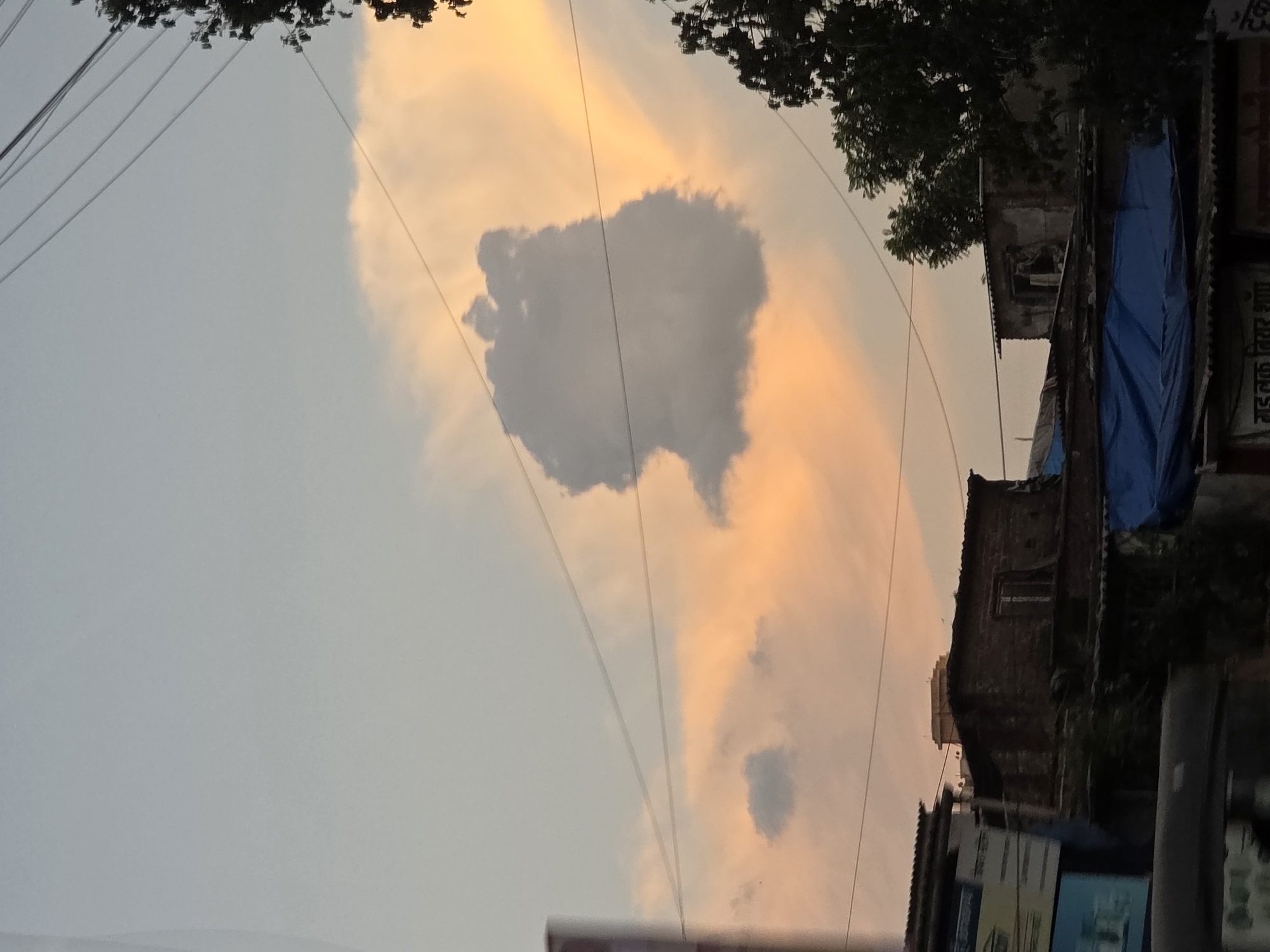 The image captures a view of a cluster of buildings in a densely populated urban area, likely a slum or a lower-income neighborhood in Mumbai, India. The sky above is a hazy mix of pale blue and warm orange, suggesting it might be sunset or sunrise. A prominent, dark cloud formation is suspended in the sky, its shape vaguely resembling an animal or a whimsical figure.
The buildings are a mix of structures with corrugated iron roofs and some with what appear to be brick walls. They are crowded together, with narrow spaces between them. One building in the foreground has a blue tarp covering part of its roof, possibly for weather protection. Another building to the left has various signs attached to its facade, some of which are partially obscured. Electrical wires crisscross the scene, a common sight in such urban settings. Dense foliage from trees is visible on the right side of the image, contrasting with the man-made structures.
No people are clearly visible in the frame. The overall impression is one of a bustling, everyday scene in a busy Indian city, with a touch of natural beauty provided by the sky and cloud formation. The lighting is soft and diffused, contributing to a somewhat muted color palette.