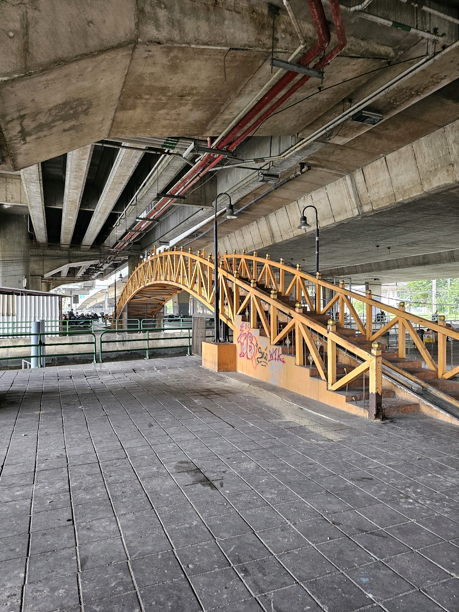 This is a photograph of a pedestrian bridge, located under a larger, elevated concrete overpass, in Bangkok, Thailand. The bridge is painted bright orange with decorative wooden railings and has stairs leading up to it. The ground in the foreground is paved with dark grey square tiles. Red and white pipes are visible running along the underside of the overpass. In the background, through the open structure of the overpass, a line of motorbikes and several people can be seen, suggesting the presence of a street or parking area. The overall scene depicts an urban infrastructure environment with elements of both public passage and utilitarian construction.