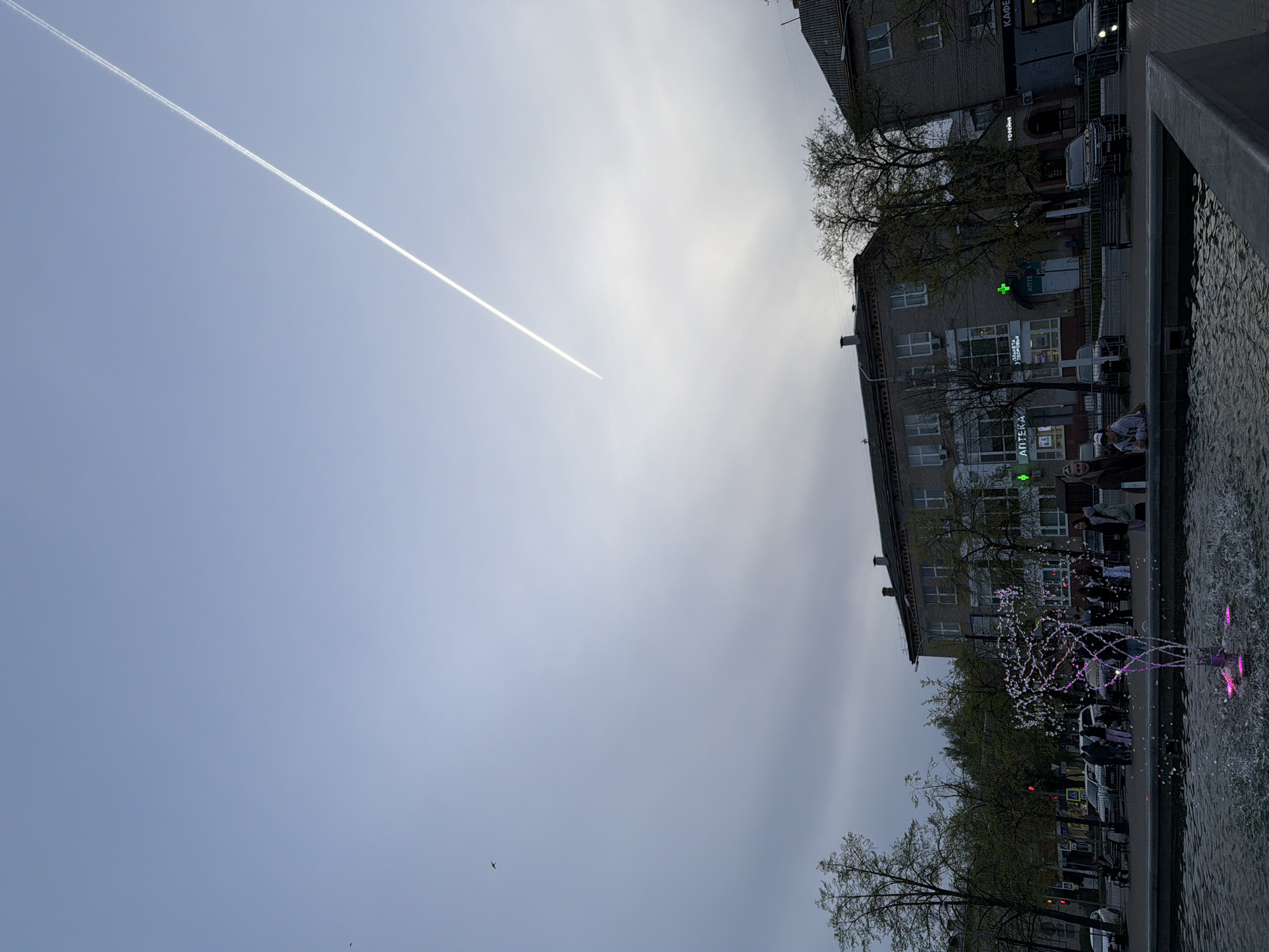 Captured from a low angle, the image showcases an urban scene in what appears to be the city of Chaikovsky, Russia. A jet contrail streaks across a predominantly overcast sky, hinting at high altitude aircraft activity.
A multi-story building occupies the right side of the frame, featuring architectural details such as regularly spaced windows and a storefront with Russian text, possibly indicating a pharmacy ("Аптека"). Pedestrians are visible in front of the building, suggesting a street-level retail area.
In the foreground, a fountain with illuminated water jets adds a touch of vibrancy. The water is slightly blurred, indicating movement, and the fountain's design incorporates pink lighting elements. The image gives the feeling of a cool overcast day in a Russian city center.
