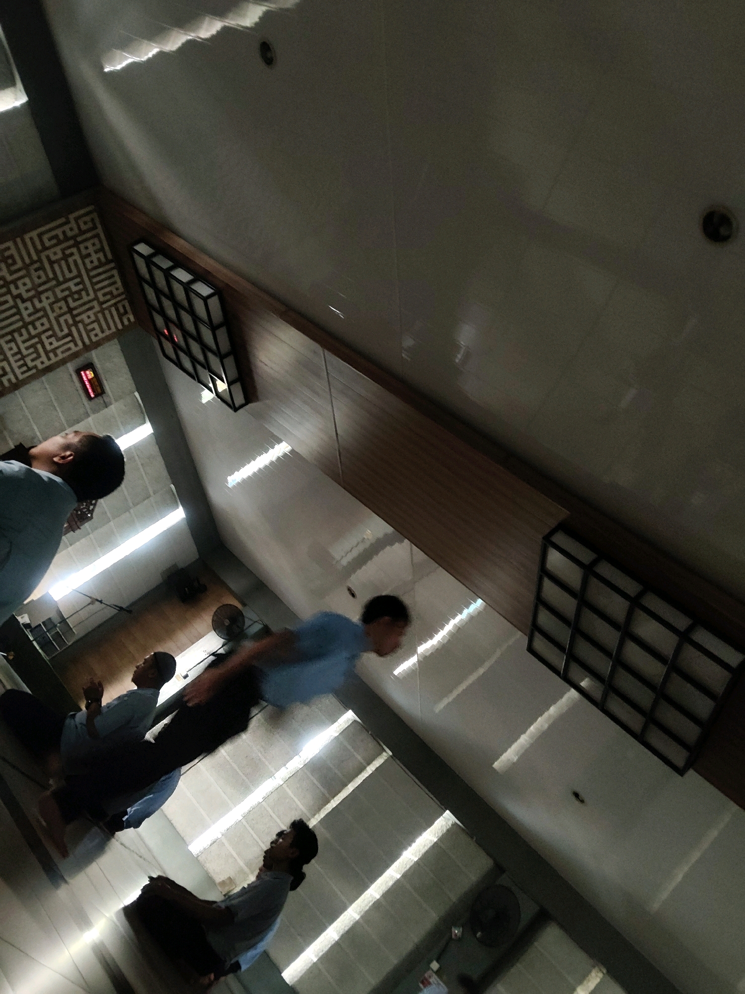 The image captures an interior scene, likely a mosque, with several young men engaged in prayer. The perspective is from above, looking down at an angle. The floor appears to be tiled, reflecting the bright overhead lights that cast long, geometric shadows.
On the left side of the image, a man in a light blue shirt and dark pants is seen from the back, facing away from the camera. His head is tilted upwards. Further back, a man in a similar light blue shirt and dark pants is also visible from the back.
In the center of the frame, a man in a light blue shirt is in motion, appearing to be in the midst of prayer or movement. His body is blurred due to the motion. Below him, another man in a light blue shirt and dark pants is kneeling, facing the camera and looking upwards, with his hands clasped.
To the right of the kneeling man, another individual in a light blue shirt and dark pants is also kneeling and looking upwards. A fan is visible on the floor behind them.
The walls are light-colored, with some decorative elements. On the upper left wall, there is a large, intricate geometric pattern in a lighter color. Above this pattern, there is a red digital display. Two black square light fixtures with a grid pattern are mounted on the wall. The ceiling is a light color with visible beams and panels, and the lighting creates distinct patterns of light and shadow. The overall impression is of a place of worship in use.
Based on the presence of Islamic prayer and the architectural style, this scene is consistent with a mosque located in Mekarmukti, Indonesia.