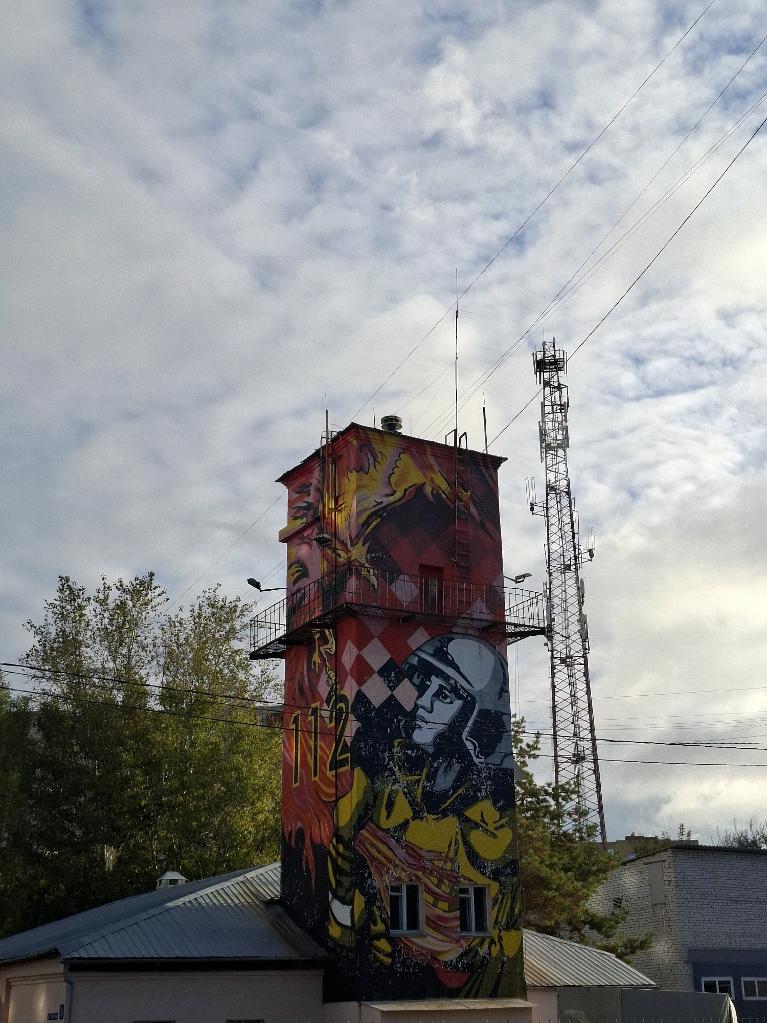 This is a photograph of a graffiti-covered tower in the city of Tyumen, Russia, with a cell tower in the background. The tower is painted with a mural depicting a person in a helmet, flames, and the number "112". The tower has a metal balcony and windows. The sky is cloudy. In the background, there is a tall metal cell tower with multiple antennas. To the left of the tower is a green tree and to the right are buildings. The image appears to be taken from a low angle, looking up.