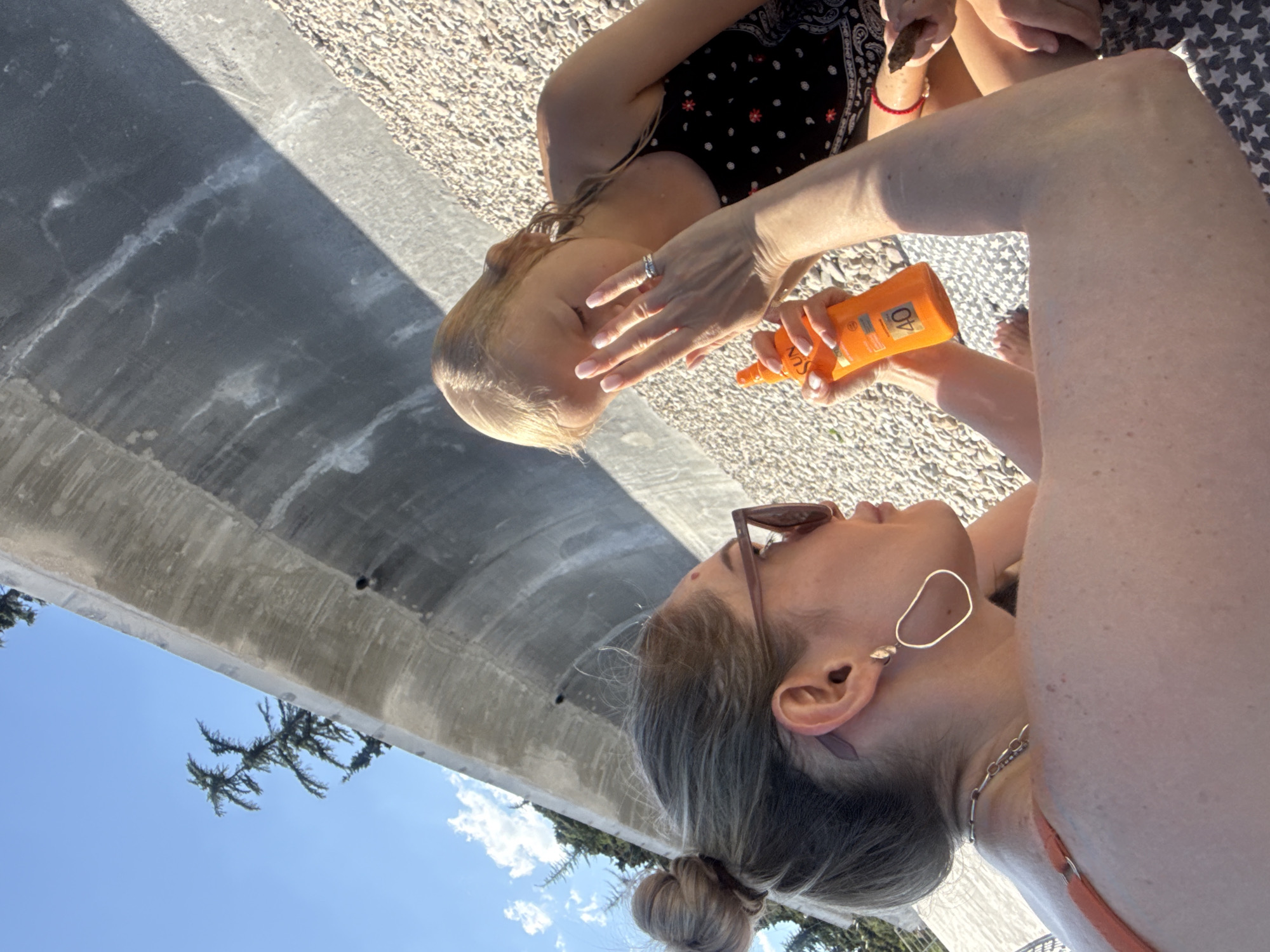 Two individuals are on a pebble beach in Utios, Ukraine, near a large, curved concrete structure. The person in the foreground, a woman with blonde hair in a bun and wearing sunglasses and an orange swimsuit, is applying sunscreen from an orange bottle with SPF 40 to the face of a young blonde girl. The girl is wearing a black and white patterned dress. The environment appears to be outdoors with a clear blue sky and trees visible in the background. The concrete structure is textured and appears to be part of a seawall or embankment.