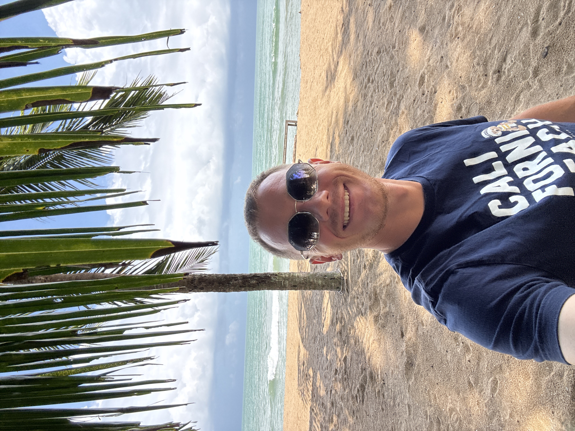 A fair-skinned man with short brown hair and a closely trimmed beard smiles as he takes a selfie on a sandy beach in Ban Khuk Khak, Thailand. He is wearing a dark blue t-shirt with white text that reads "CALI FORNI BEACH" and a small image of a bear wearing a sailor hat. He is also wearing aviator sunglasses with a silver frame. In the background, palm fronds hang overhead, casting shadows on the sand. Beyond the beach, the turquoise ocean meets a cloudy sky. A single palm tree stands near the center of the frame, behind the man. A wooden bench is visible in the distance on the beach.