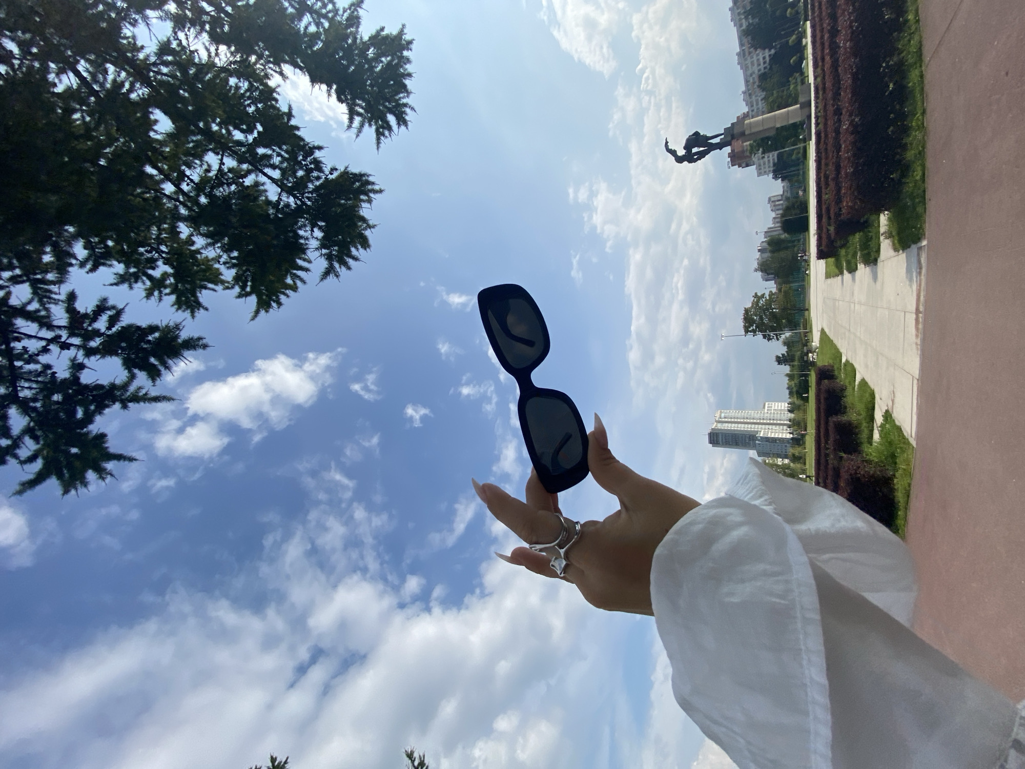 In an outdoor, urban setting, a person's hand, adorned with silver rings and elongated fingernails, holds up a pair of black sunglasses against a bright blue sky filled with white, scattered clouds. The arm and shoulder of the person are visible, clad in a white garment. In the background, a park-like area features manicured bushes, paved walkways, and distant city buildings, including a tall, modern skyscraper. A bronze statue of two figures is prominent on a pedestal. Evergreen tree branches extend into the frame from the top of the image. The sun appears to be overhead, casting a bright light. The image is captured from a low angle, looking upwards.