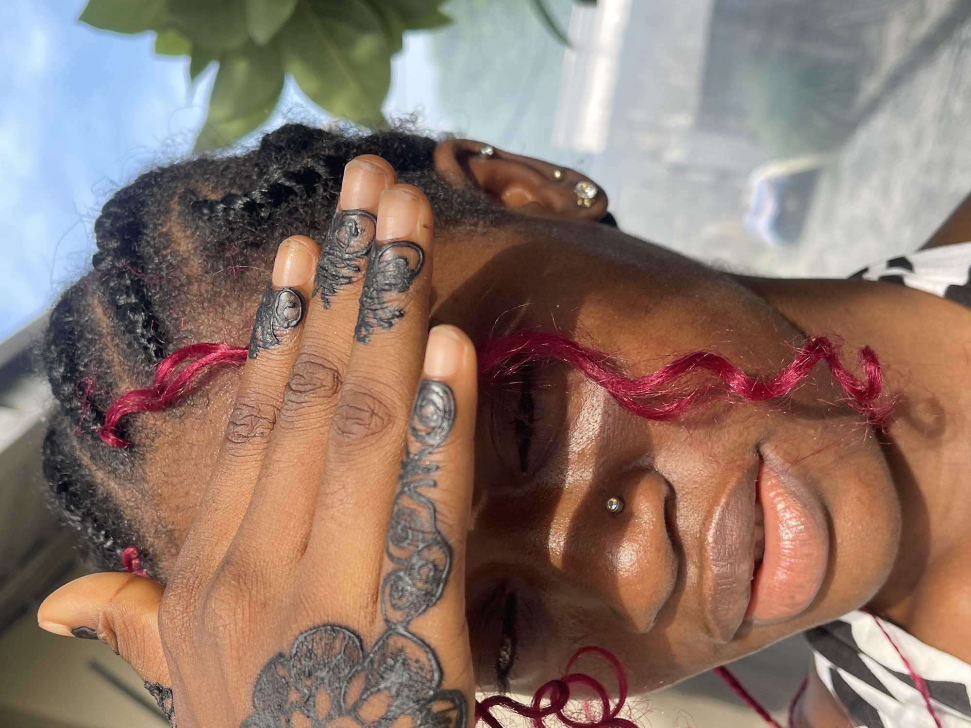 The media file captures a close-up, angled portrait of a person, likely female, with a dark skin tone, possibly taken in Ado, Nigeria. The individual is positioned with their head tilted, and one hand, adorned with intricate black henna-like designs on the fingers and back, partially covers their face.
The person has dark, braided hair with distinctive strands of vibrant reddish-pink hair woven throughout, adding a pop of color. Multiple piercings are visible: a silver stud on the nostril and several small, sparkling stud earrings in the earlobe and cartilage. The lips are full, and part of an eye is visible beneath the hand. A glimpse of a black and white striped garment can be seen at the bottom right of the frame.
The background is softly blurred, revealing a light blue sky and green foliage, suggesting an outdoor setting or an indoor space with a large window. The presence of bright, natural light indicates the image was taken during the daytime. No text is visible in the image. The overall mood appears relaxed or contemplative, though specific emotions are obscured by the pose.