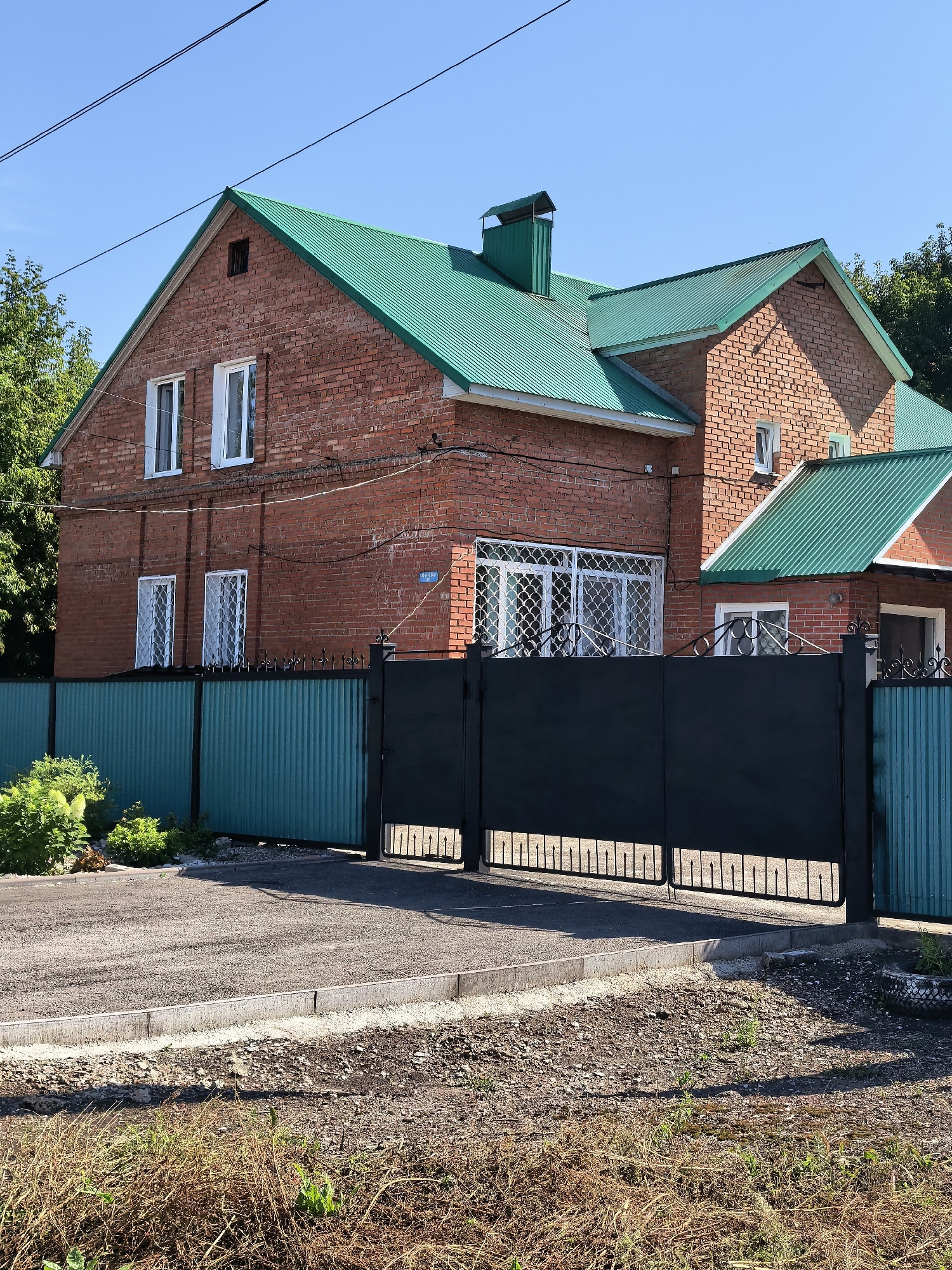 The image shows a two-story brick house with a green metal roof, situated behind a black metal gate and a teal corrugated metal fence. The house has several white-framed windows, some with decorative metal grilles. A chimney with a green cap is visible on the roof. The property is surrounded by green foliage and trees. A paved driveway leads up to the gate. The ground in the foreground is covered with dry grass and gravel. The sky is clear and blue. This is a residential property located in Sterlitamak, Russia.