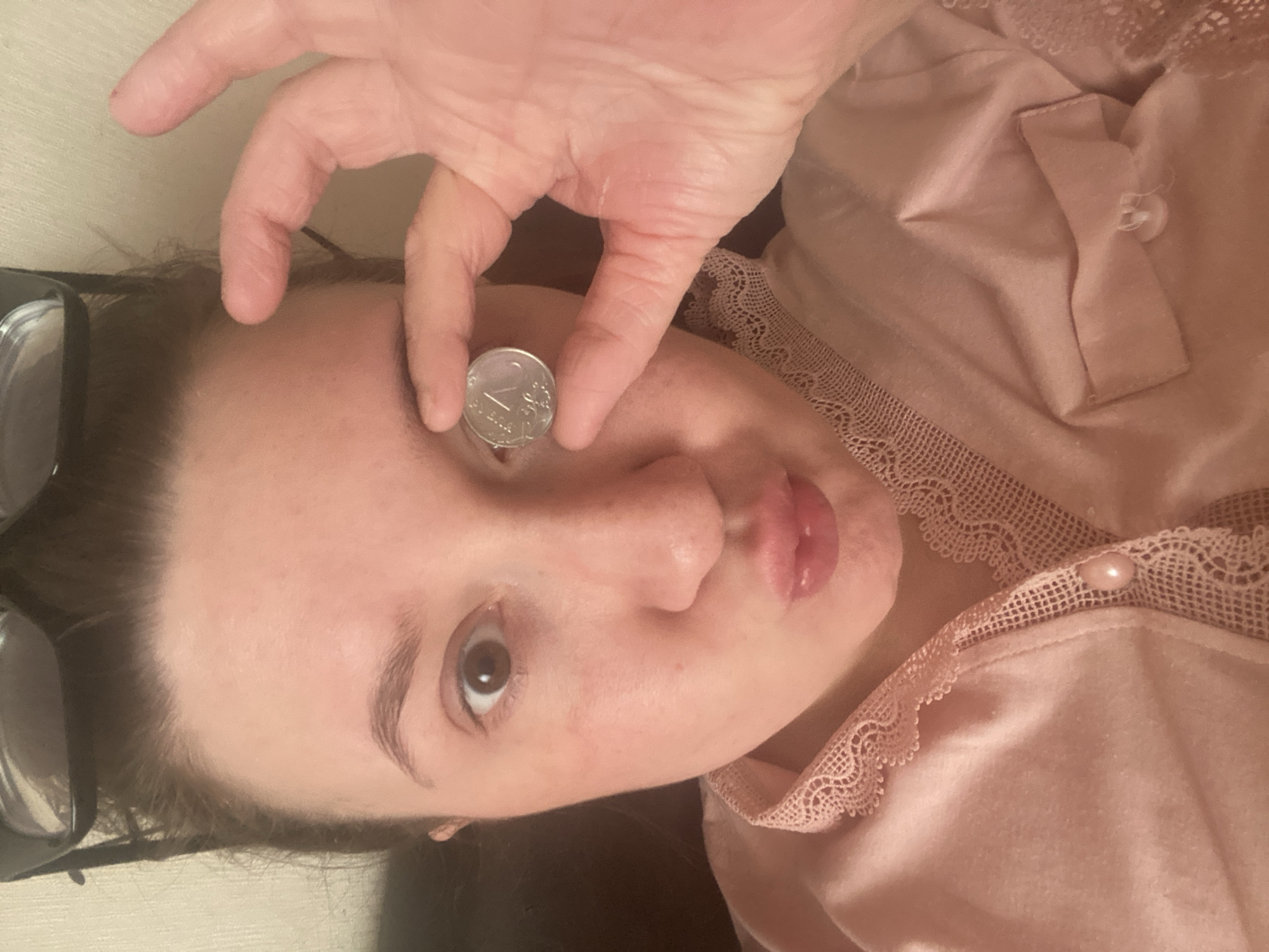 In Chaikovsky, Russia, a close-up, high-angle selfie photograph depicts an adult female indoors. Her brown hair is pulled back, with dark-framed eyeglasses resting on her forehead. She has light skin, brown eyes, and her lips are slightly puckered. Her left hand holds a silver-colored Russian 1 Ruble coin between the thumb and index finger, positioned directly in front of her right eye, obscuring it. Her left eye remains open and focused on the camera. She wears a light pink garment, possibly satin or silk, with intricate lace detailing along the collar and a single pale, circular button visible on the front opening. The background consists of a plain, light-colored wall or surface, indicating an indoor private setting.
