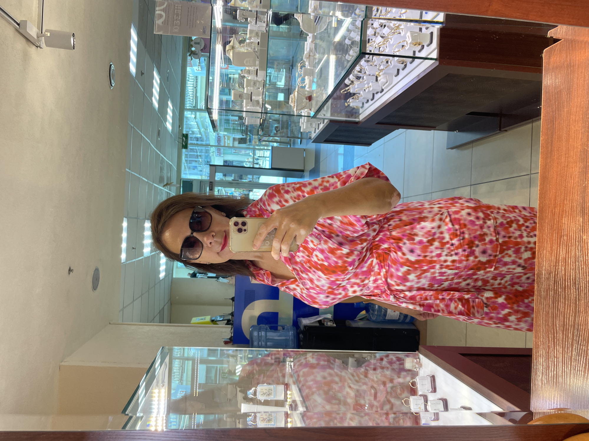 A woman takes a mirror selfie in a jewelry store. She is wearing a pink and red patterned dress and sunglasses. She is holding a gold iPhone in her right hand. In the background, there are display cases filled with jewelry and a water cooler. The store appears to be a modern retail environment with bright overhead lighting. The scene is indoors. The signage in the background suggests the location is in Sochi, Russia, as indicated by the Cyrillic text that appears to read "Ювелирные изделия" (Jewelry).