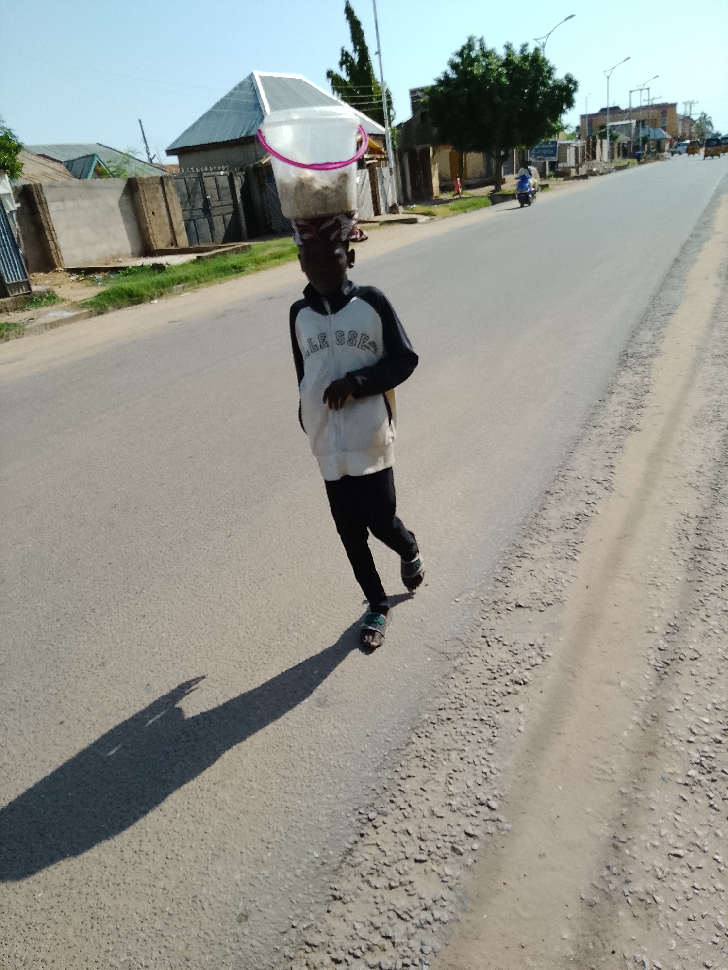 In Wauro Leggal, Nigeria, a young person walks down a paved road carrying a bucket of grain on their head. The individual is wearing a white and black jacket and black pants. The road is bordered by a dusty shoulder and appears to be in a residential area with buildings and trees visible in the background. The sky is clear and blue, suggesting a sunny day. The person's shadow stretches long across the road, indicating the sun is low in the sky.