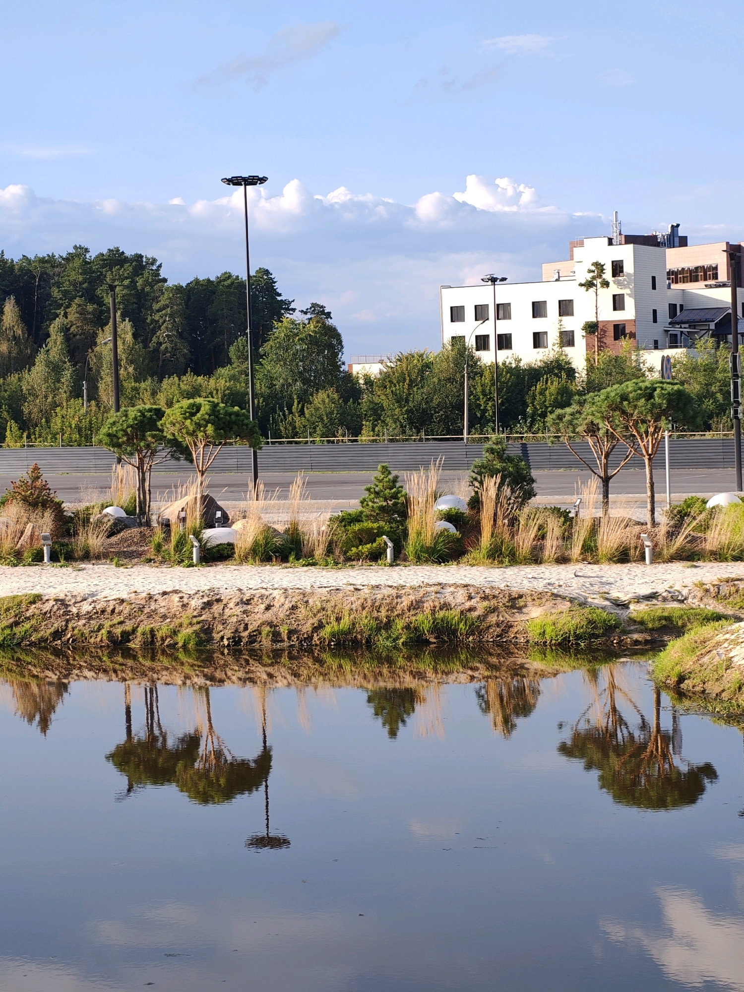 The image depicts an outdoor scene with a body of water in the foreground, reflecting the sky and surrounding landscape. The water is calm, with ripples disturbing the perfect reflection. The bank of the water features sandy soil and green grass.
Behind the water is a landscaped area with several small, ornamental trees, some of which appear to be pines, interspersed with tall, dry grasses and large white spherical objects that could be decorative elements. There are also some rocks and small white lights along the pathway.
Further in the background, a metal fence runs horizontally across the frame, separating the landscaped area from a road. Beyond the fence, a dense line of trees forms a natural backdrop. In the upper right portion of the image, a modern, multi-story building with a white facade and numerous dark windows is visible. The sky is blue with scattered white clouds. Two tall, dark lampposts with multiple light fixtures are prominent in the scene. One is positioned in the middle ground, and another is further to the left.
This scene is located in the "Krasnotsvet" park in Yekaterinburg, Russia, specifically in the area near the Uktus ski resort. The modern building in the background is part of the "Ural Scientific and Educational Center" complex.