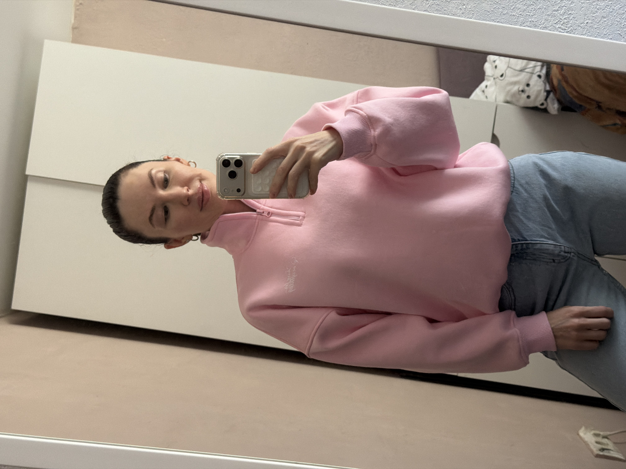 A young woman with dark hair pulled back into a bun is reflected in a mirror. She is holding a smartphone with a clear case and three camera lenses in her right hand, presumably taking a selfie. She is wearing a pink, long-sleeved, half-zip sweatshirt and light blue jeans. The reflection shows part of a white cabinet behind her and a glimpse of a patterned pillow and a brown object in the upper right corner. The room appears to be indoors with light-colored walls. There is no specific information in the image to indicate that it was taken in Perm, Russia.