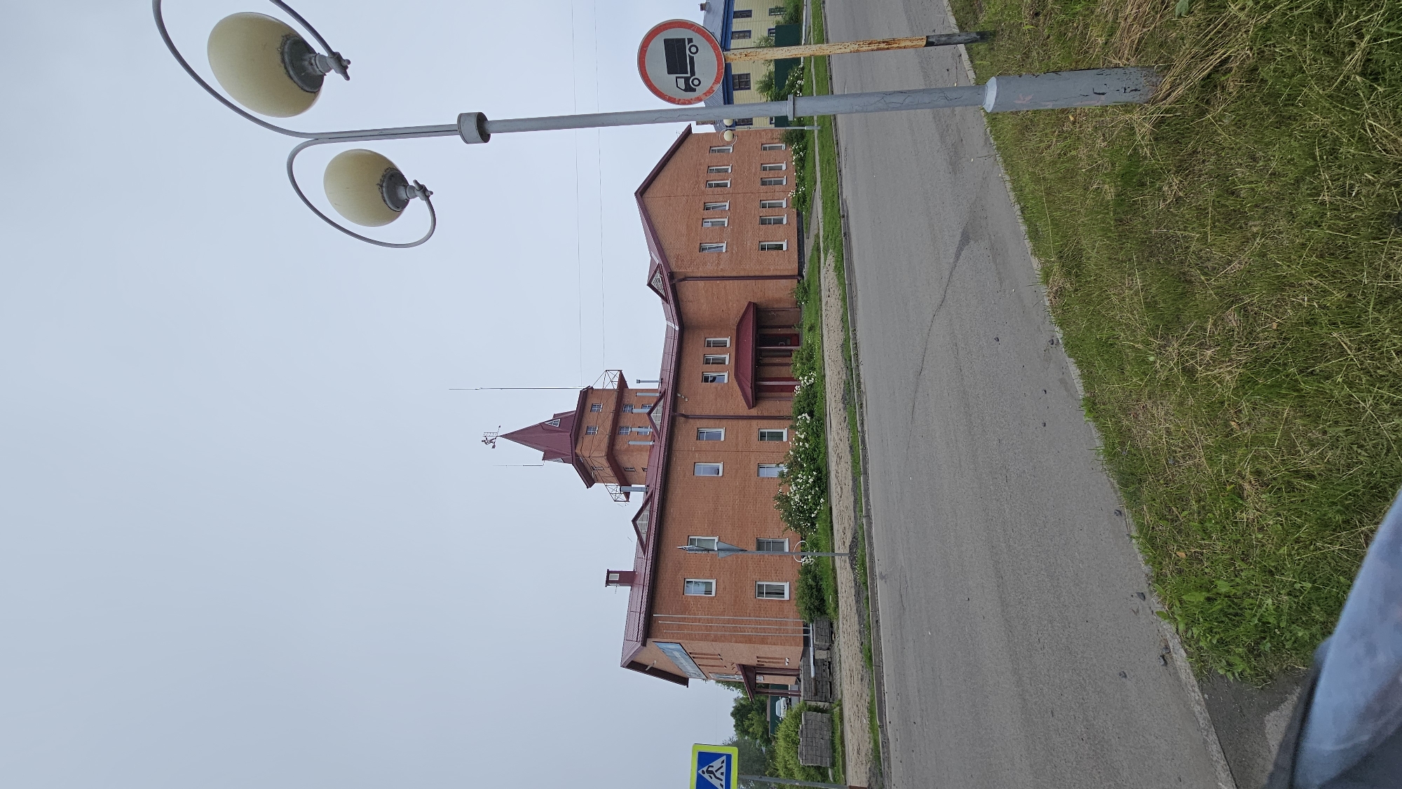 A brick building with a red roof and a central tower stands on a cloudy day. The building appears to be two stories with numerous windows. In front of the building, a road with a sidewalk is visible, lined with grass. A lamppost with two lights stands to the right of the image. A traffic sign showing a truck with a red circle and a diagonal line through it is also present. Another traffic sign depicting a pedestrian is located to the left of the building. Some building materials are stacked on the ground near the sidewalk.