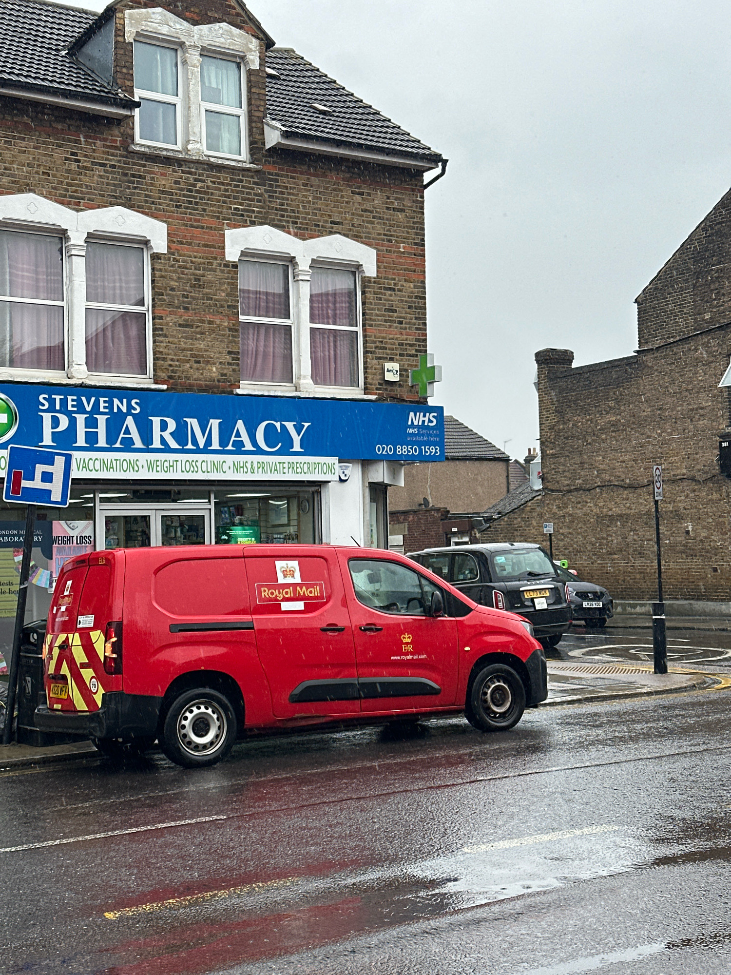 A red Royal Mail van is parked on the right side of the street, its rear facing the camera. The van is wet, suggesting it has been raining. The street is also wet and reflects the van and surrounding buildings. In the background, a pharmacy called "STEVENS PHARMACY" is visible, with a blue sign and windows displaying products. A black car is parked behind the Royal Mail van. Further in the background, there are brick buildings and a cloudy sky. The scene appears to be a street in a residential or commercial area of London, United Kingdom, on a rainy day.