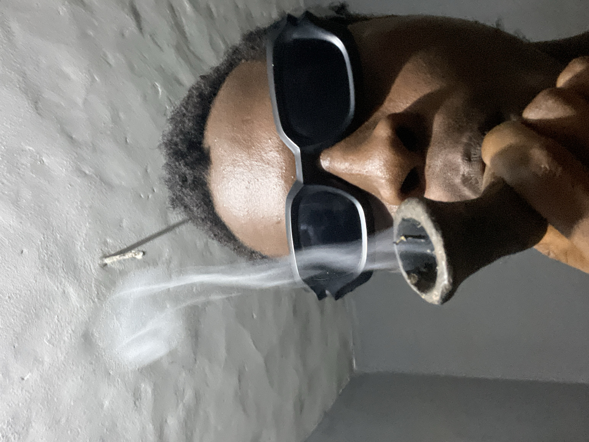 This close-up portrait, taken in Onitsha, Nigeria, features an individual with dark skin and dark, curly hair, viewed from the chest up. The person is wearing black sunglasses with silver frames, which obscure their eyes. The image is oriented vertically, with the subject's head tilted to their left.
The central activity depicts the person smoking. They are holding a dark, cylindrical object, likely a pipe or similar smoking device, to their mouth. A distinct plume of white smoke billows from the end of the device, extending towards the upper left of the frame.
The setting appears to be indoors or in a dimly lit space. The background consists of a textured, light gray wall, possibly concrete or plaster, with an uneven, slightly rough surface. A small, thin, light-colored stick or wire is visible protruding horizontally from the wall in the upper left quadrant. The lighting is subdued, suggesting evening, night, or an interior with limited illumination. No text is visible in the image. The person's expression is not discernible due to the sunglasses.