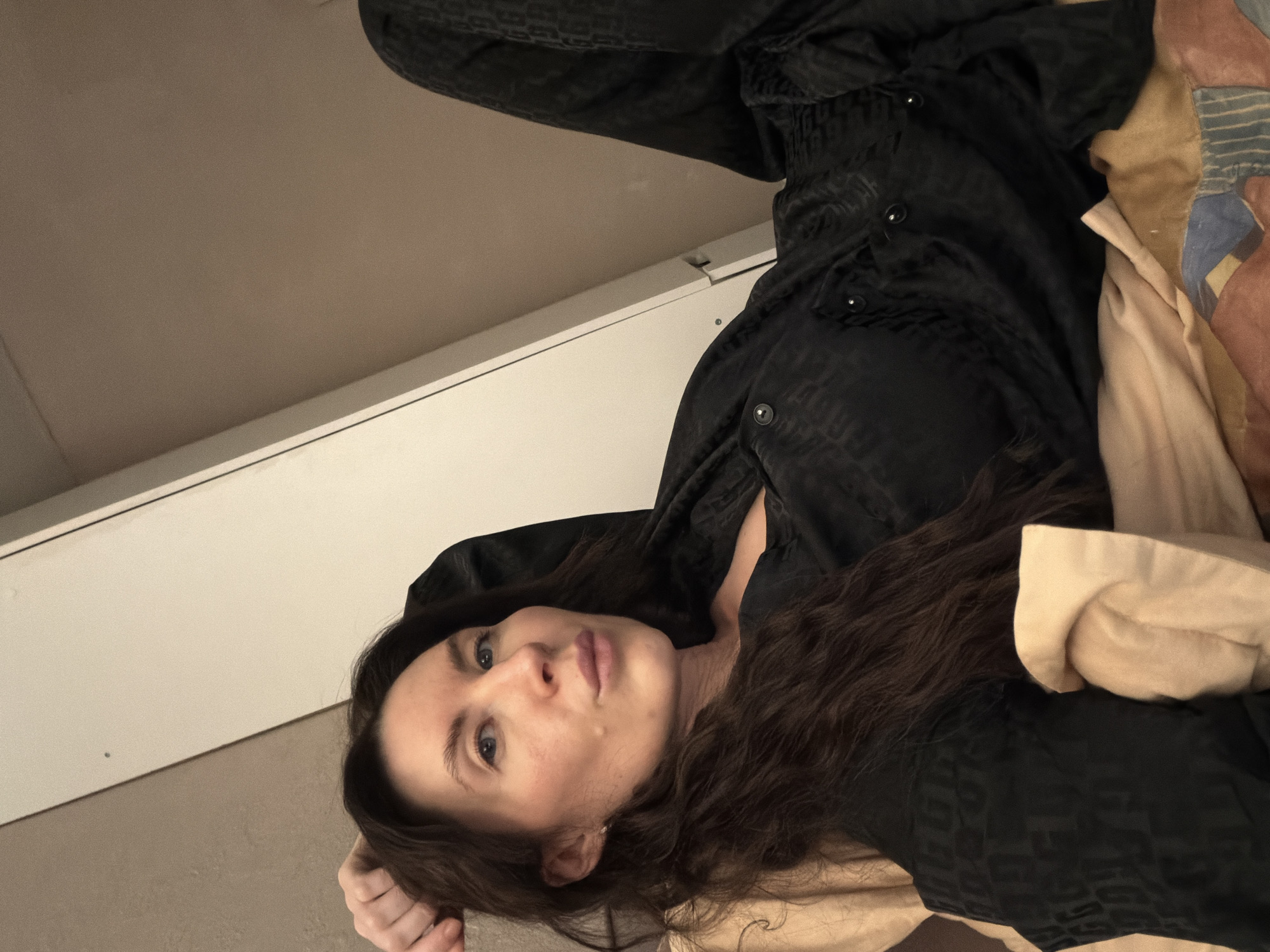 The image depicts an adult female subject positioned indoors, resting on what appears to be a bed or lounge area. The subject is oriented diagonally from the lower left to the upper right of the frame, looking directly towards the camera.
**Subject Details:**
- **Individual:** One female.
- **Hair:** Long, wavy, dark brown.
- **Facial Features:** Fair complexion, light blue eyes, a mole present on the right cheek. The subject's lips are slightly pursed.
- **Clothing:** Dressed in black pajamas with a subtle, dark-on-dark geometric or patterned fabric. The top features visible black buttons down the front.
- **Posture:** The subject is lying on her back, with her head slightly elevated and her right hand placed behind or under her head. Her body is relaxed against the bedding.
**Environment Details:**
- **Setting:** An interior room, likely a bedroom.
- **Bedding:** The subject is resting on a light beige or peach-colored pillow or cushion. Below her, a patterned blanket or sheet with segments of beige, blue, and brown is visible.
- **Background:** Behind the subject's head and extending to the upper left, a plain, light brown/beige wall is visible. To the right and upper center, a white or off-white piece of furniture, possibly a wardrobe or cabinet, is angled into the frame. This furniture item shows a vertical seam or door edge and a small dark circular mark (potentially a screw or imperfection) near its upper left edge.
- **Lighting:** The scene is moderately illuminated, consistent with an indoor environment.
**Location:**
The image provides no identifiable landmarks, signage, or distinctive architectural features to confirm a specific location within the city of Perm, Russia. The setting appears to be a generic private indoor space.