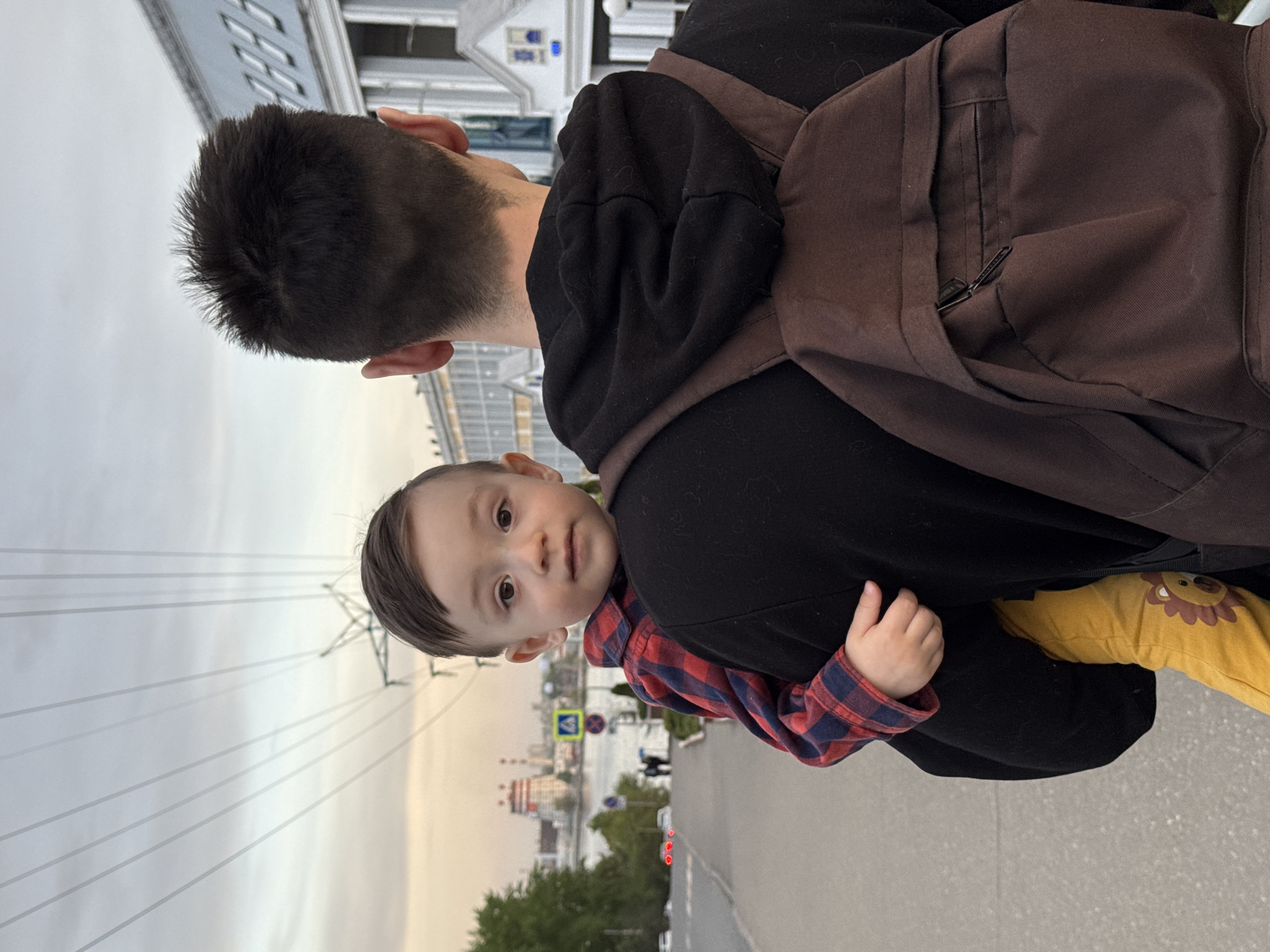 A man is carrying a young child on his back. The man has short, dark hair, and is wearing a black hoodie and a brown backpack. The child is wearing a red and blue plaid shirt and yellow pants with a lion design. The child is looking at the camera. The picture was taken on the street near the Izh river, across from the Svyato-Mikhailovsky Cathedral. The background includes city buildings, trees, power lines, and a bridge with some cars visible. The street is paved, and the sky is overcast.