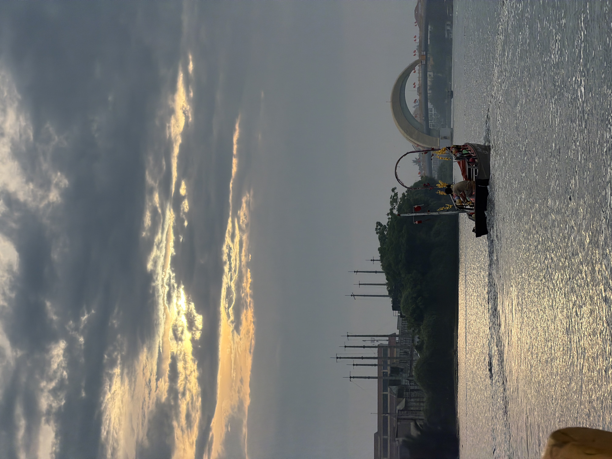 The image captures a picturesque scene over a body of water in Đà Nẵng, Vietnam, likely during sunrise or sunset. The sky is dramatic, filled with dark grey clouds interspersed with brilliant golden sunlight breaking through, creating striking vertical rays of light.
On the left side of the frame, a traditional-style boat is visible on the water. The boat is adorned with yellow and red decorations, including an elaborate arched structure, possibly a ceremonial canopy. At least one person in light-colored attire is on the boat, bent over as if engaged in an activity like preparing equipment or rowing. The water itself is calm but shimmering, reflecting the golden light from the sky in streaks across its surface.
The distant bank of the water features lush green foliage and various structures. A prominent, light-colored arched bridge serves as a distinctive landmark, spanning the water or connecting parts of the land. Small red flags are visible lining the distant bank and possibly along the bridge structure. Further inland, industrial-looking poles or masts and other buildings can be seen, partially obscured by the trees. The overall atmosphere is serene, emphasizing the beauty of the natural light and the city's features. No visible text is present in the image.