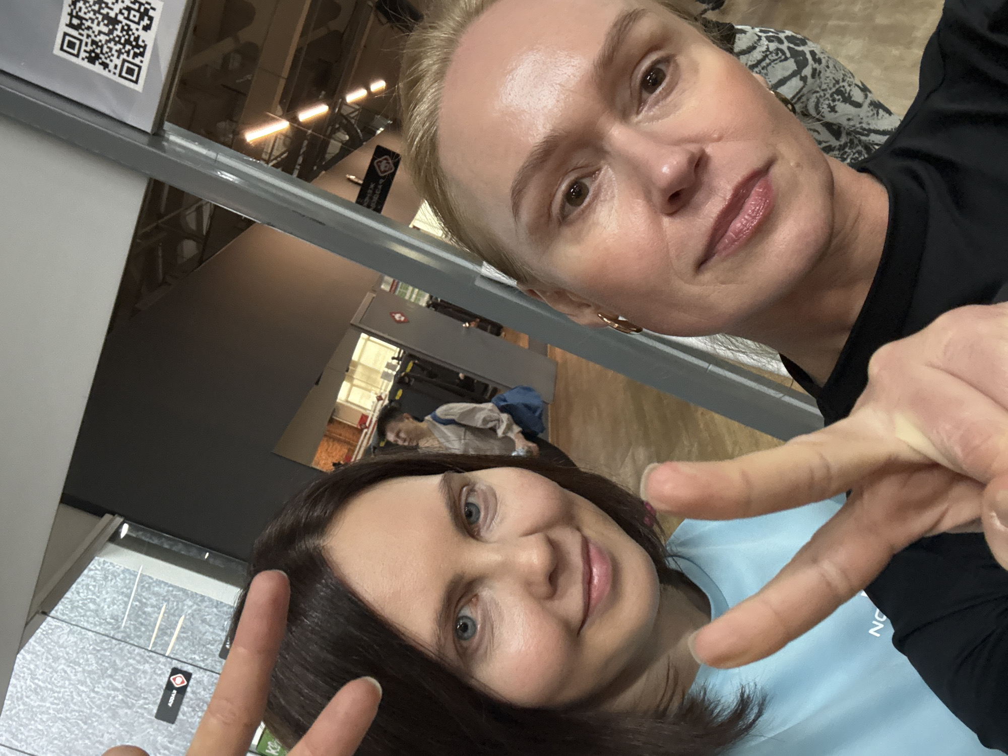 Two women are taking a selfie indoors. The woman at the top has short, blonde hair and is wearing a black shirt. The woman at the bottom has shoulder-length brown hair and is wearing a light blue shirt. Both are making peace signs with their fingers. Behind them, there is an interior space that appears to be a hallway or waiting area with fluorescent lighting and various signage on the walls. In the background, there are people sitting in the waiting area. Based on the small sign visible in the frame, this photo was taken in Chaikovsky, Russia. A QR code is visible on a sign in the upper left corner.