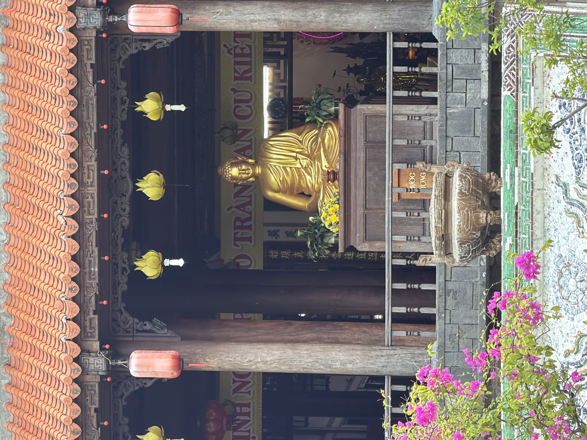 The image depicts a golden Buddha statue seated on a wooden pedestal within a temple in Nha Trang, Vietnam. The statue is adorned with yellow flowers and surrounded by greenery. In front of the Buddha is an ornate incense burner. Red lanterns and yellow lotus-shaped lanterns hang from the temple's eaves. The temple features a traditional tiled roof and wooden pillars. Pink bougainvillea flowers cascade in the foreground, adding a vibrant touch to the scene. A banner with Vietnamese text is visible behind the Buddha statue. The overall atmosphere is serene and spiritual.