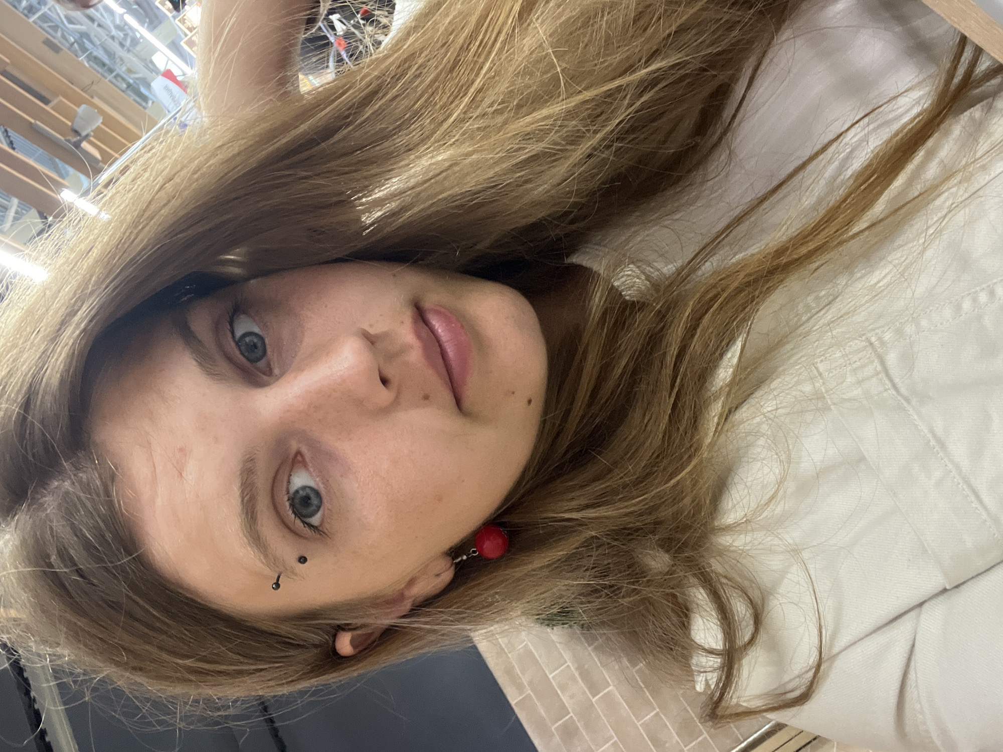 The image is a close-up, indoor selfie of a young person with long, blonde hair. The subject has light blue eyes, fair skin with freckles, and is wearing a white collared shirt. They have an eyebrow piercing with two small black studs and a dangling red earring on their left ear. The background is blurred, but appears to be an indoor commercial space with visible ceiling structures and lighting, possibly a retail environment. There is no discernible indication of the location being Minsk, Belarus.