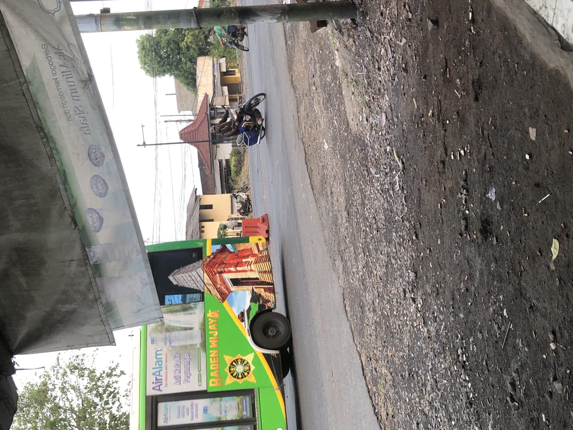 In Kludan, Indonesia, a bus emblazoned with "Air Alam" and "RADEN WIJAYA" is parked on the side of a road, its green exterior featuring images of a waterfall and a temple. In the background, several buildings with reddish-brown roofs are visible, along with trees and a utility pole. A man on a motorcycle drives past the bus, while another man on a motorcycle is further down the road. The road itself is paved and cracked, with a debris-strewn dirt shoulder.