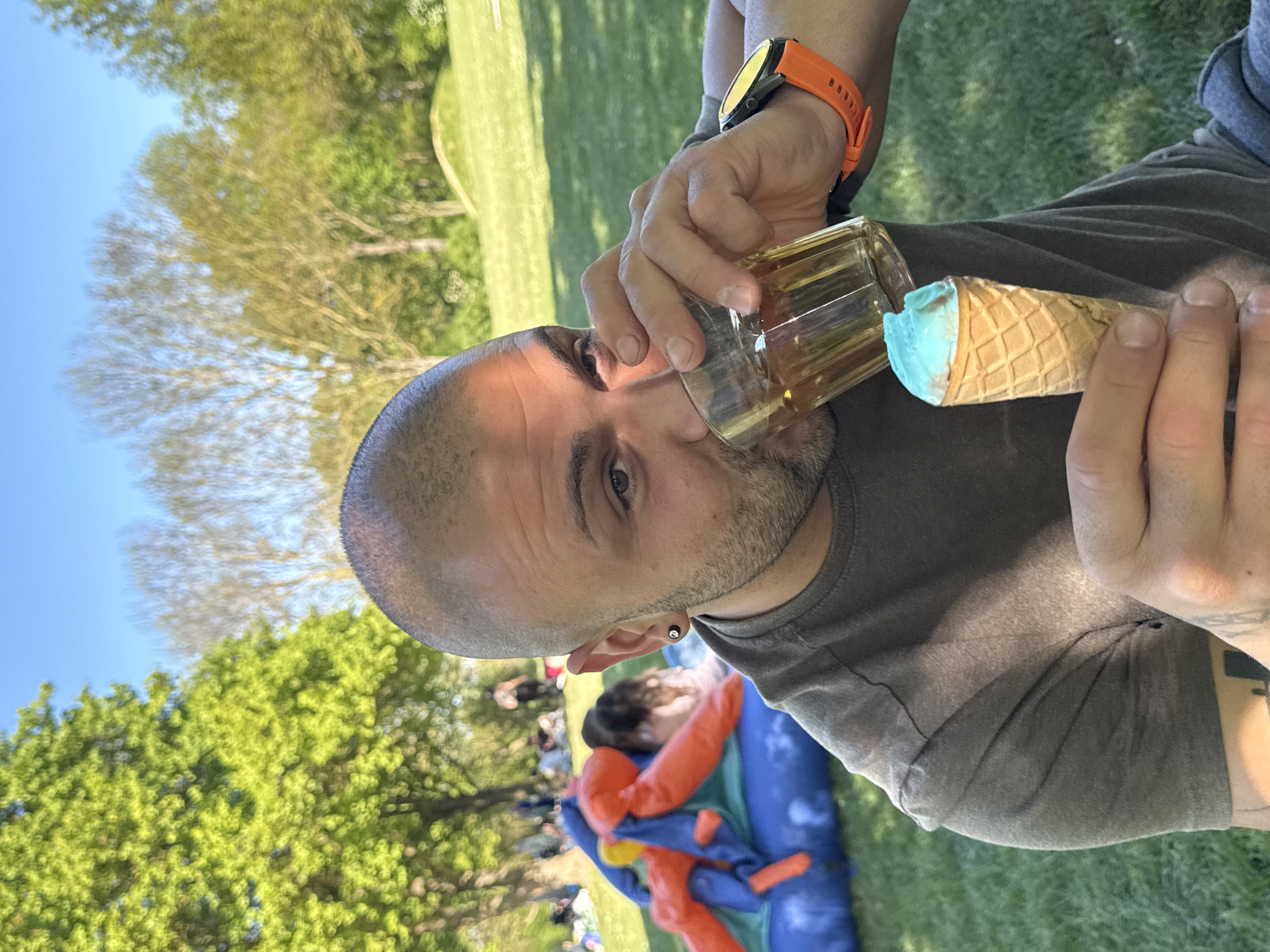 The image displays a man in the foreground drinking from a clear glass, while simultaneously holding an ice cream cone with vibrant blue ice cream in his right hand. He has a shaved head and a short beard, and is wearing a dark grey t-shirt along with an orange smartwatch on his left wrist. An earring is visible in his left ear.
The setting is outdoors on a sunny day, with a clear blue sky and lush green trees and grass in the background, suggesting a park or open field. In the mid-ground, blurred figures, including children, are visible around a large blue and orange inflatable structure, possibly a bouncy castle or play area, indicating a lively social gathering or event. The bright natural light suggests it is daytime. The overall atmosphere appears relaxed and festive, taking place in Ditzingen, Germany. There is no visible text in the image.