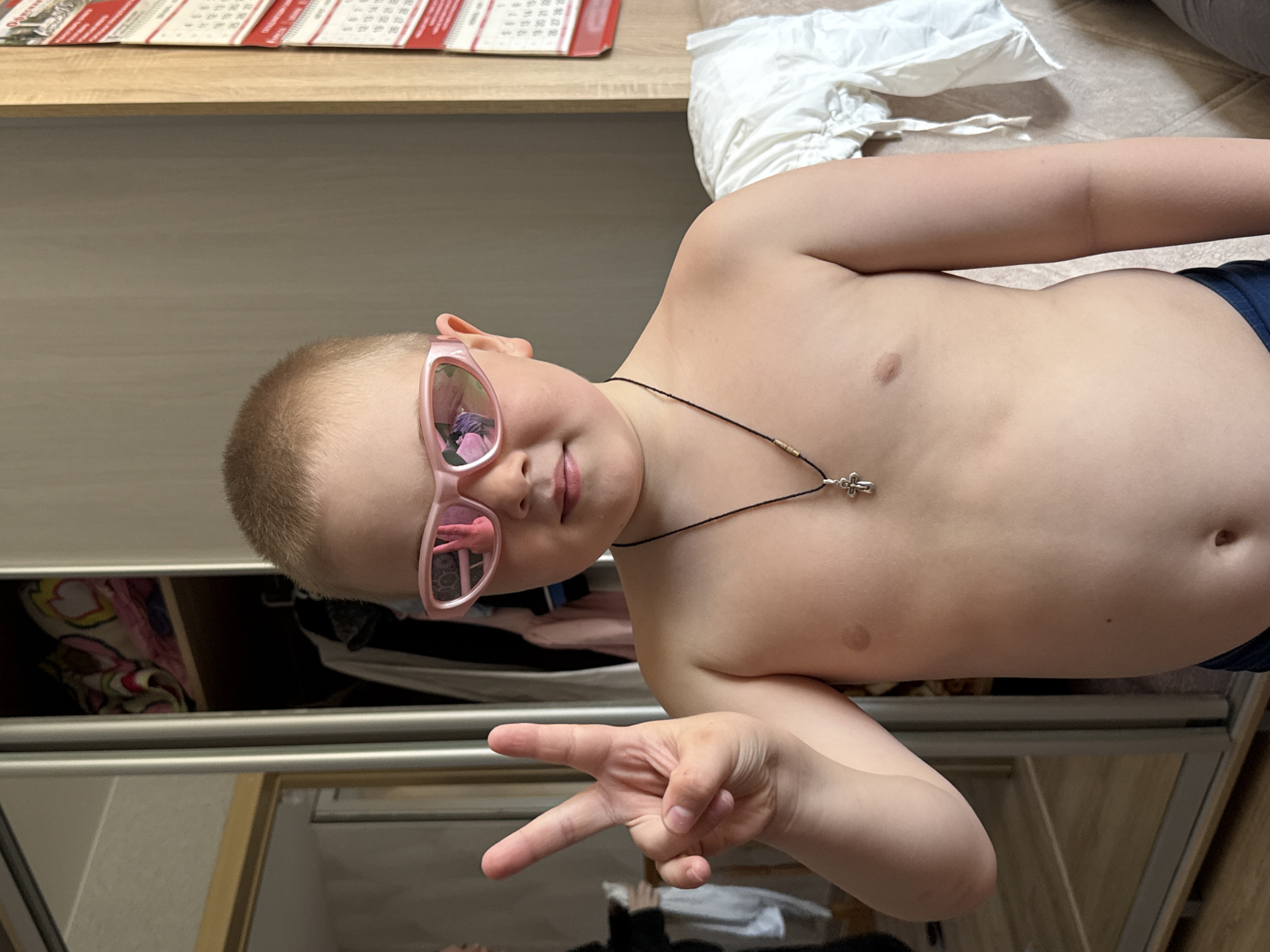 A fair-skinned child, likely male, with a buzz cut, is lying down, wearing pink sunglasses and a necklace with a cross pendant. The child is giving the peace sign with their right hand. The background consists of a closet with a mirrored door, bedding, and a calendar. The location is likely an interior space, possibly a home, in Zhigalki, Russia.