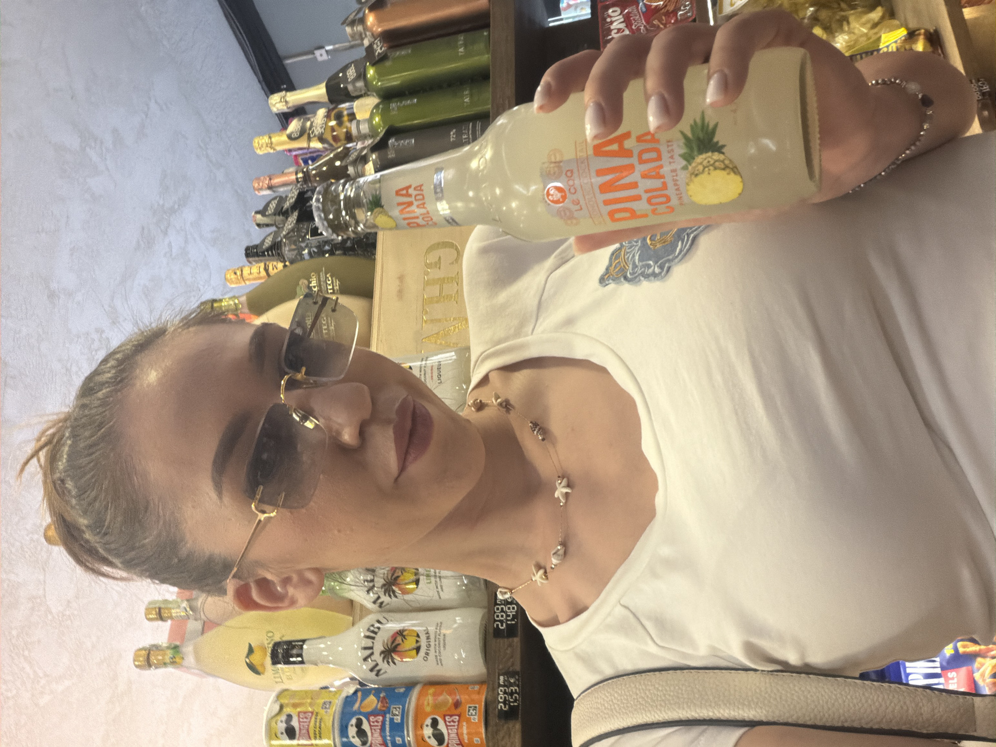 A woman with her hair tied up in a bun and wearing gold-rimmed sunglasses holds up a bottle of "Pina Colada" beverage. She is wearing a white t-shirt and a shell necklace. In the background, there are shelves stocked with various bottles of alcohol and snacks. The scene appears to be a retail environment, possibly a liquor store or a market. This image was taken in Sofia, Bulgaria.