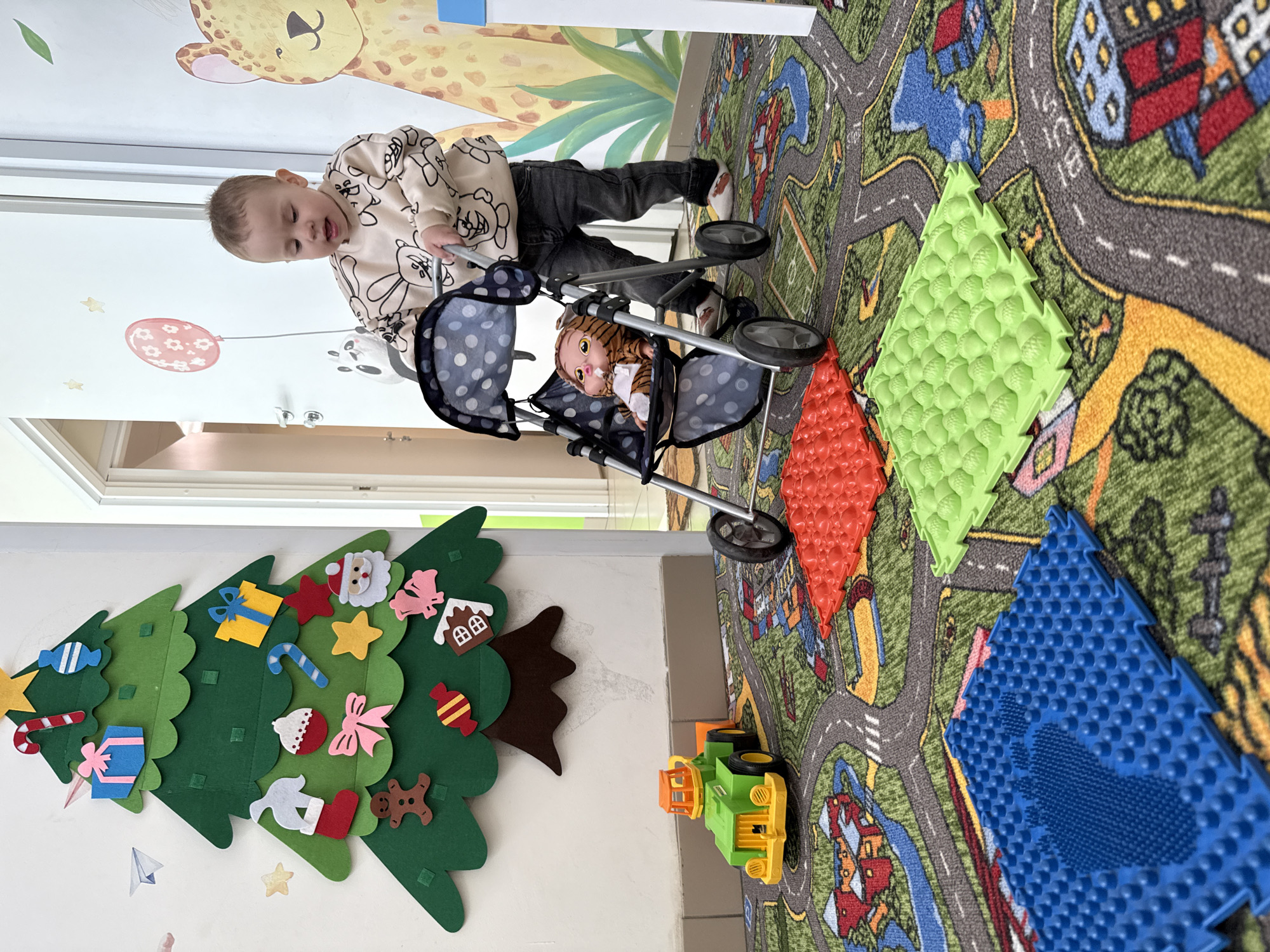 A toddler wearing a long-sleeved shirt with cartoon character drawings and dark-colored pants is pushing a toy stroller with a doll inside. The toddler is on a patterned rug resembling a city street map. On the rug are three textured foam mats in red, green, and blue. A toy truck is also on the rug. A felt Christmas tree is mounted on the wall. The background includes a wall with a giraffe cartoon and a white wall with a window. The scene is in Rusanovo, Russia.