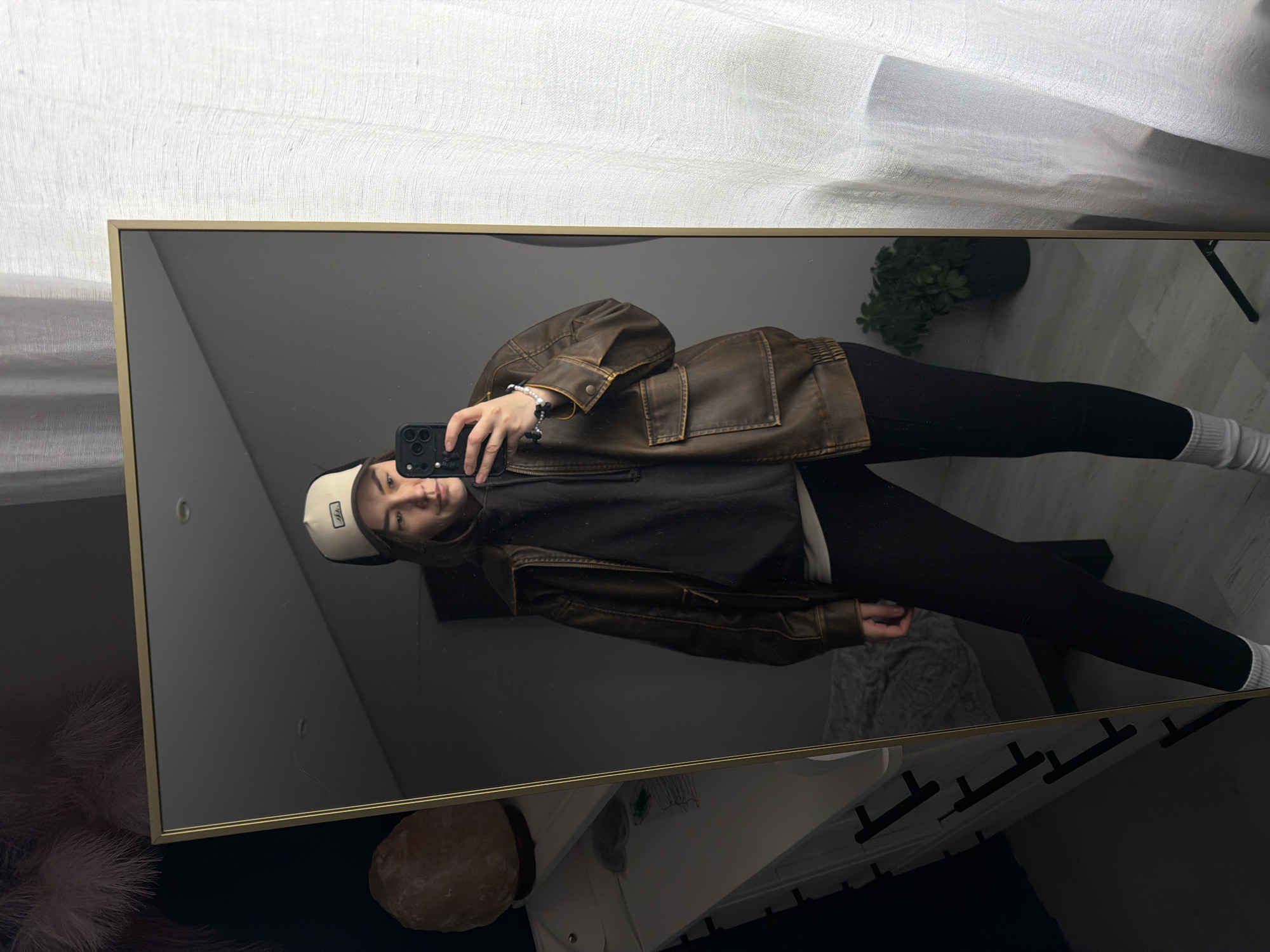 A person is reflected in a gold-framed mirror. The person is wearing a tan and black baseball cap, a black sweatshirt, a brown leather jacket, black leggings, and white socks. The person is holding a smartphone and taking a photo of themself. The background of the mirror shows a white curtain, a green plant in a black pot, a white dresser, a pink feathered object, and a pink rock. The location is unknown and there is no evidence to suggest it is in Chaikovsky, Russia.