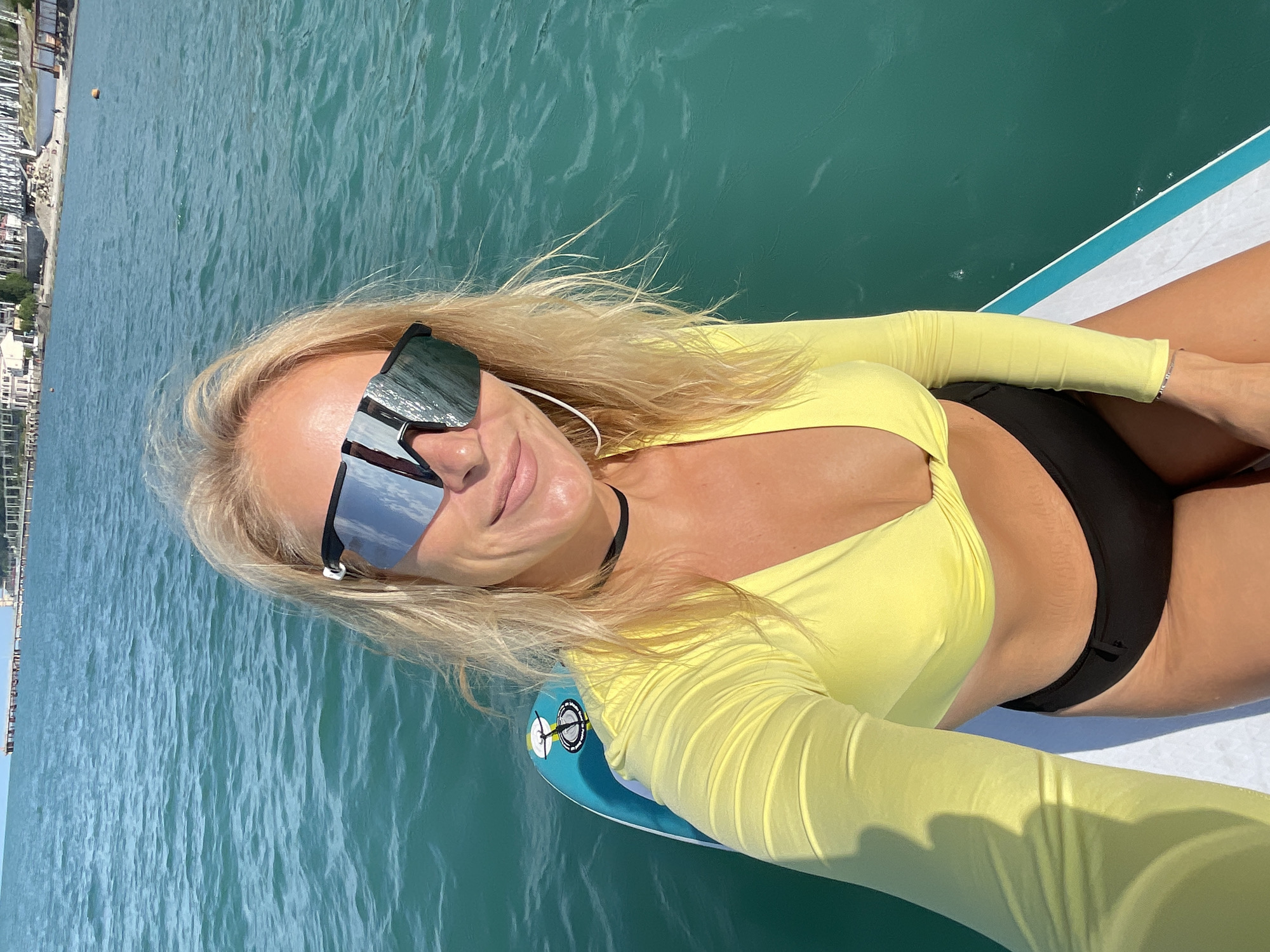 A woman with blonde hair and sunglasses is pictured on a paddleboard in the Black Sea, near the city of Sochi, Russia. She is wearing a yellow long-sleeved bikini top and black bikini bottoms. The water is turquoise and choppy, with a distant shoreline visible.