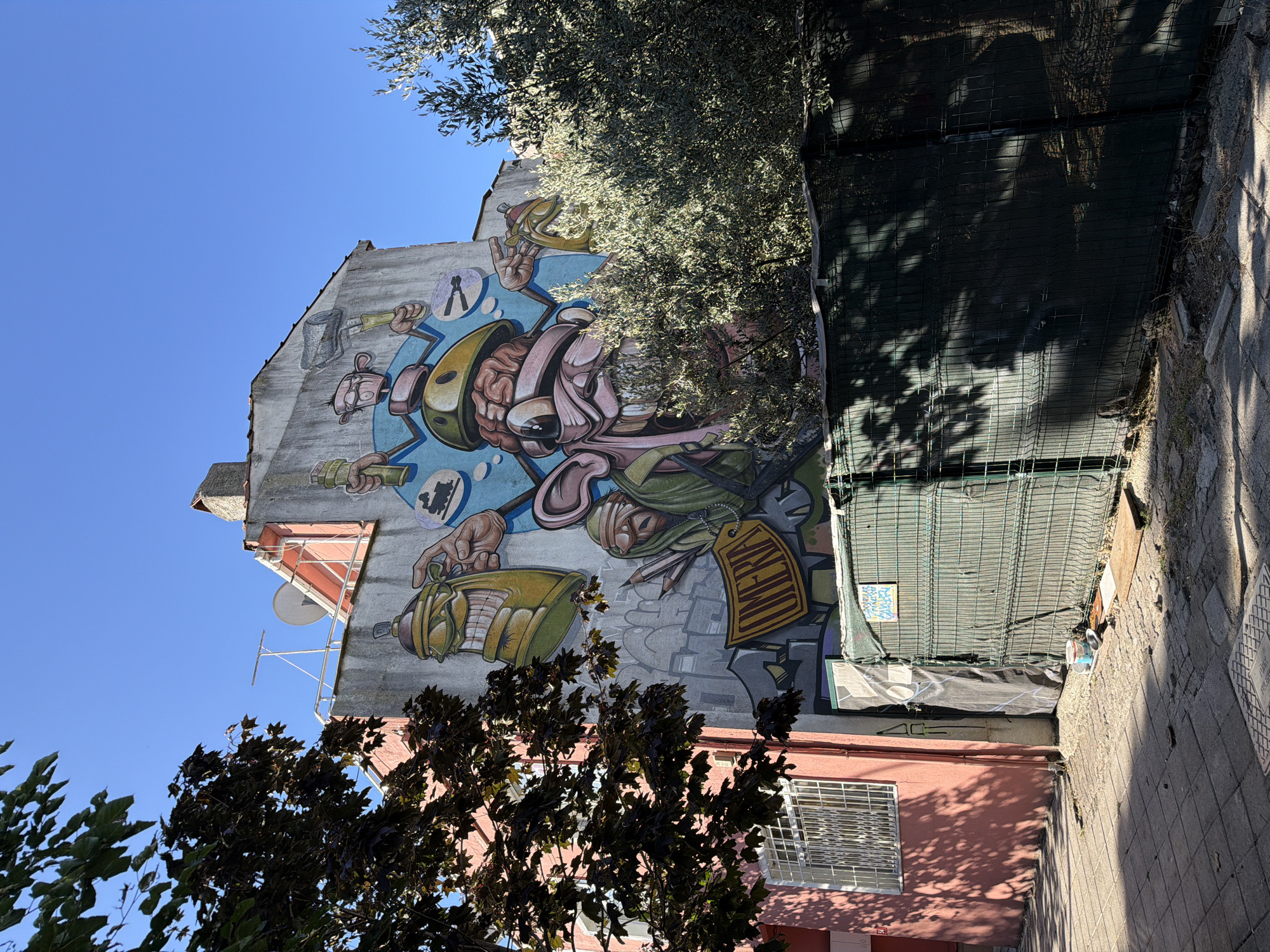 The image shows a colorful mural painted on the side of a building in Kadıköy, Turkey. The mural depicts a cartoonish character with exaggerated features, holding spray cans and a paint roller, surrounded by elements that suggest street art and creativity. The building itself is multi-story with a pink facade on the left side and a grey, textured wall on the right where the mural is located. A clear blue sky is visible above. In the foreground, there is a sidewalk with a cobblestone pattern, a green fence, and some vegetation, including a dark-leaved tree on the left and an olive tree on the right. Shadows from the trees are cast across the sidewalk and fence, indicating sunlight. Part of the fence has a green mesh netting. There is a satellite dish and antenna on the roof of the building.