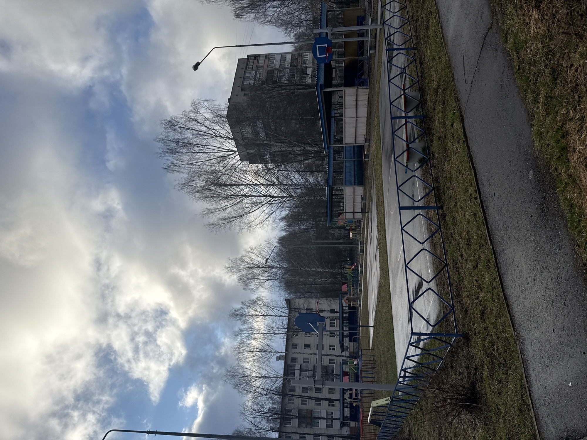The image depicts an outdoor urban scene, likely a residential area or park in Chaikovsky, Russia, on what appears to be an overcast day. The perspective is from ground level, looking towards a series of multi-story buildings and a recreational space.
The sky dominates the upper portion of the frame, filled with a dramatic interplay of bright white, gray, and darker clouds, with hints of blue sky peeking through, suggesting the sun is either obscured or low in the sky.
In the mid-ground, several bare deciduous trees with intricate branch patterns stand tall, silhouetted against the sky and partially obscuring the apartment blocks behind them. These buildings are typical Soviet-era residential structures, with multiple windows and visible balconies, appearing somewhat weathered.
In the foreground and middle ground, a recreational area is visible, featuring a blue metal fence with diamond patterns that runs horizontally across the image. Beyond this fence is an asphalt surface, possibly a playground or a basketball court, with a blue basketball hoop and backboard clearly standing out. Further in the distance, hints of other colorful playground equipment can be seen.
To the right, an asphalt path or road runs parallel to the fence, bordered by sparse, brownish-green grass. Another streetlight with a curved arm is visible near the buildings.
No people are present, and no specific activity is taking place, giving the scene a quiet, somewhat somber atmosphere. The bare trees and muted color palette suggest late autumn, winter, or early spring. There is no visible text in the image.