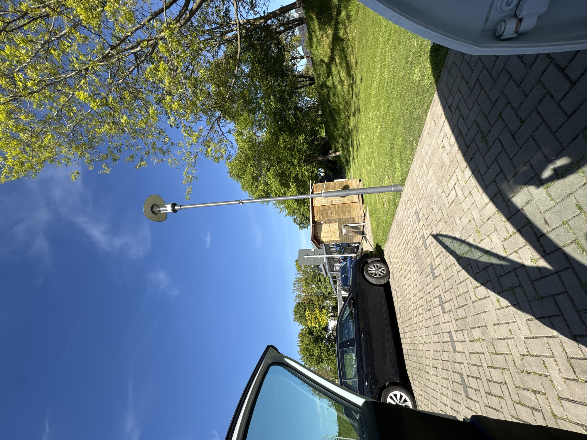 The image shows an outdoor scene, likely in Wassenberg, Germany, due to the prompt's indication. A car, seemingly a black sedan, is parked on a brick-paved surface, possibly a driveway or parking area. The car door is open, partially obscuring the view. A lamppost stands nearby, casting a shadow on the paving. In the background, there is a small wooden shed and a grassy area with trees. The sky is blue with some scattered clouds, suggesting a clear day.