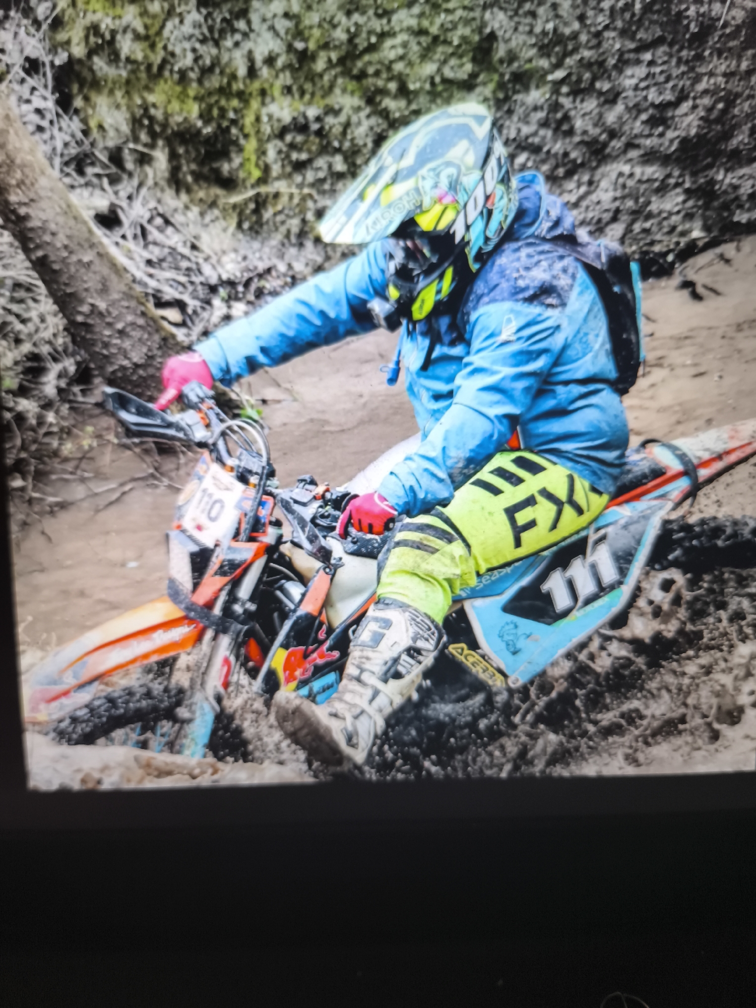 A person is riding a motocross bike in a muddy, wooded area. The rider is wearing a blue jacket, yellow and black pants with "FX" on the side, a helmet with colorful designs, pink gloves, and white boots. The motocross bike is orange and blue with the number "110" on the front and "111" on the side. The environment appears to be a forest with a muddy ground, indicating the bike is struggling through the thick mud. A tree trunk is visible in the background. The location is not specified as Enina, Bulgaria.
