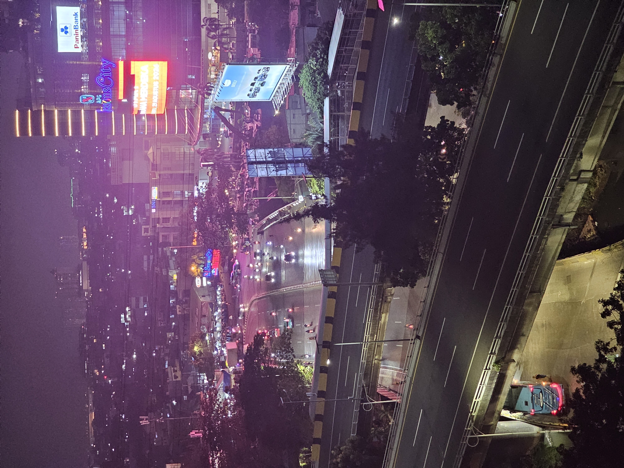 The image captures an elevated nighttime view of a bustling intersection in West Jakarta, Indonesia, likely near the Seasons City mall. A multi-lane highway, wet from recent rain, is illuminated by streetlights and the glow of city buildings. Vehicles, including cars and numerous motorcycles, navigate the roadway, their headlights and taillights creating streaks of light.
In the background, the Seasons City complex is prominently visible with its towering facade adorned with bright neon signage, including "SeasonsCity" and "PaninBank." Another large billboard featuring "GEELY" vehicles is also visible. The surrounding urban landscape is a tapestry of buildings and streetlights, indicating a densely populated and active metropolitan area. A blue bus is parked under a concrete overpass, its brake lights illuminated. A single person stands near the bus. Trees and foliage are scattered throughout the scene, softening the urban concrete. The overall atmosphere is dynamic and vibrant, characteristic of a busy city at night.
