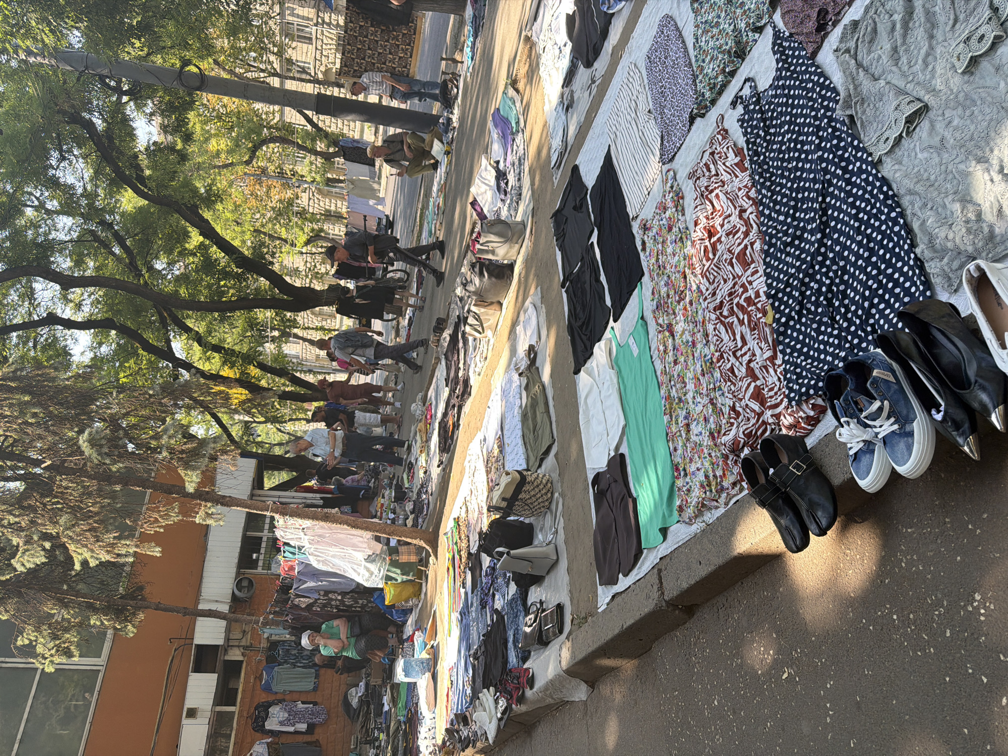 The image depicts an outdoor flea market scene, likely in Chișinău, Moldova, given the context. Numerous stalls are set up along a sidewalk, displaying a wide assortment of clothing and accessories laid out on white sheets and tarps. People are browsing the merchandise, with some individuals actively engaged in shopping and others standing around. The environment suggests a warm, sunny day, with trees providing shade over the market area. The background shows buildings and a street, indicating an urban setting.