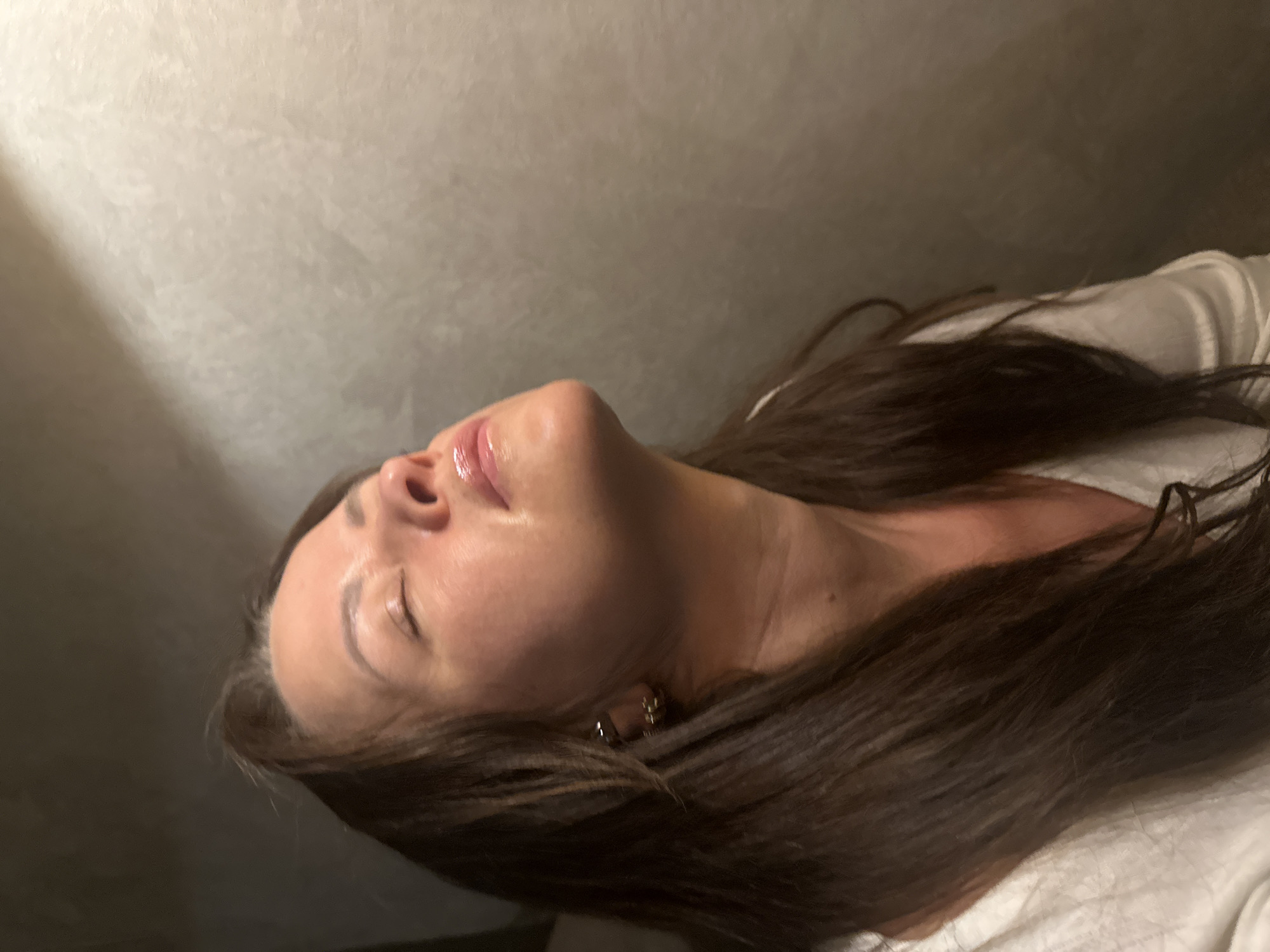 An overhead shot depicts a woman lying down indoors in Chaikovsky, Russia, with her head tilted upwards. Her long, dark brown hair cascades around her, partially covering her shoulders and a light-colored top, possibly white or cream. Her eyes are closed or nearly closed, and her lips are slightly parted, appearing glossy or moisturized. Her skin looks smooth and clear. A small, golden-toned earring is visible on her left earlobe. The background consists of a plain, light-colored wall, which shows some texture and subtle shadows, indicating an indoor setting with directed lighting. The overall impression is one of calmness or relaxation, possibly during a moment of rest or a beauty treatment. No text is visible in the image.