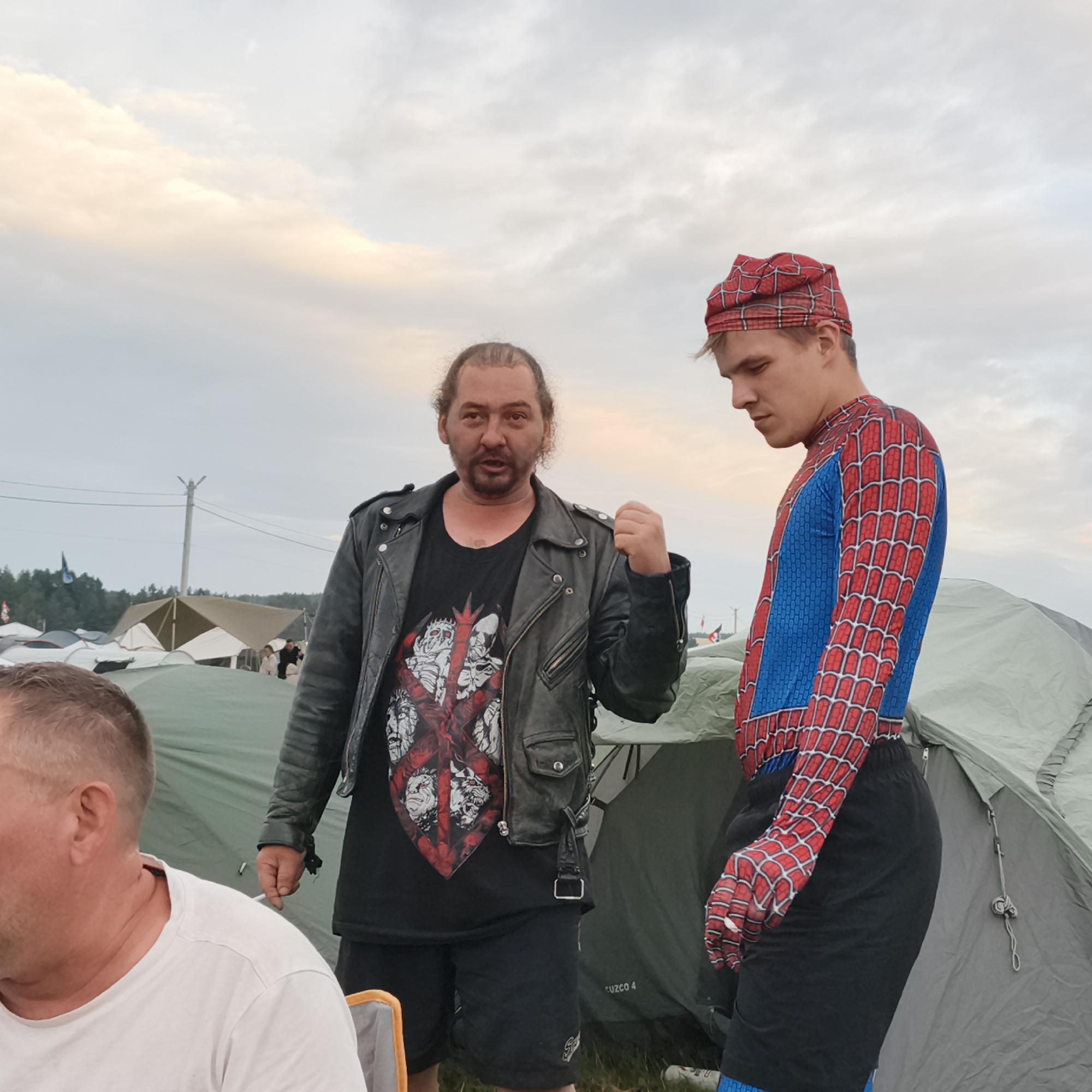 In Lagunovo, Russia, a man in a black leather jacket and a graphic t-shirt stands with his fist clenched, looking towards the camera. Beside him, a young person in a Spider-Man costume with a matching bandana stares downwards. They are positioned in an outdoor setting, possibly a campsite, with tents visible in the background under a cloudy sky. Another person, seen from behind, is seated in the foreground.