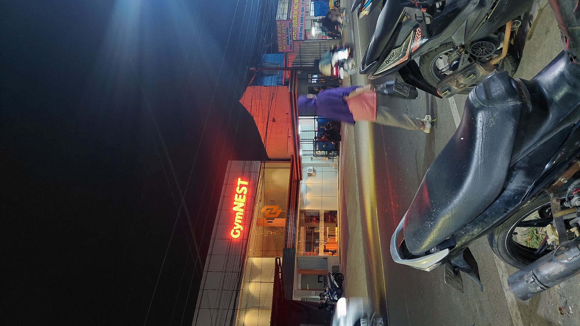 A nighttime outdoor scene in Bantarsoka, Indonesia, captures a gym named "GymNEST" illuminated by bright red neon signage. The gym, located on a street, has large glass windows revealing the interior. In the foreground, two motorcycles are parked on the roadside, one in sharp focus and the other blurred. A person wearing a purple headscarf and pink shirt is walking past the motorcycles, blurred by motion. Other vehicles and figures are visible in the background, also appearing blurred due to the low light and movement. The street is lit by streetlights and the glow from the businesses, creating a dynamic and somewhat hazy atmosphere.