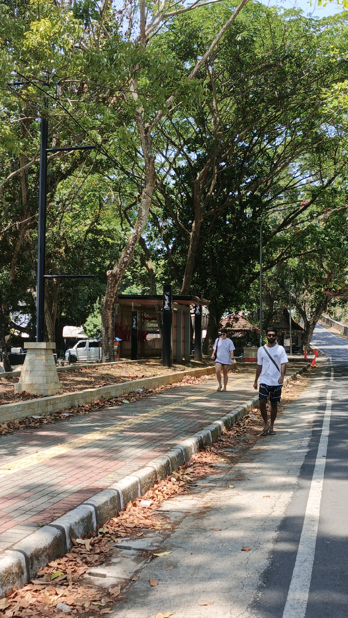 Two individuals are walking along a paved pathway in Desa Batukaras, Indonesia. The pathway is bordered by a stone curb and dry leaves. To the left of the pathway, there is a dirt area with scattered debris and a white van. Further back, a small covered structure with open sides is visible. The scene is surrounded by lush green trees providing ample shade. A paved road runs alongside the pathway on the right. The sky is clear and bright, indicating a sunny day. The man in the foreground is wearing a white t-shirt and black shorts, with a dark bag slung across his shoulder. The woman walking ahead of him is wearing a white t-shirt and grey shorts.