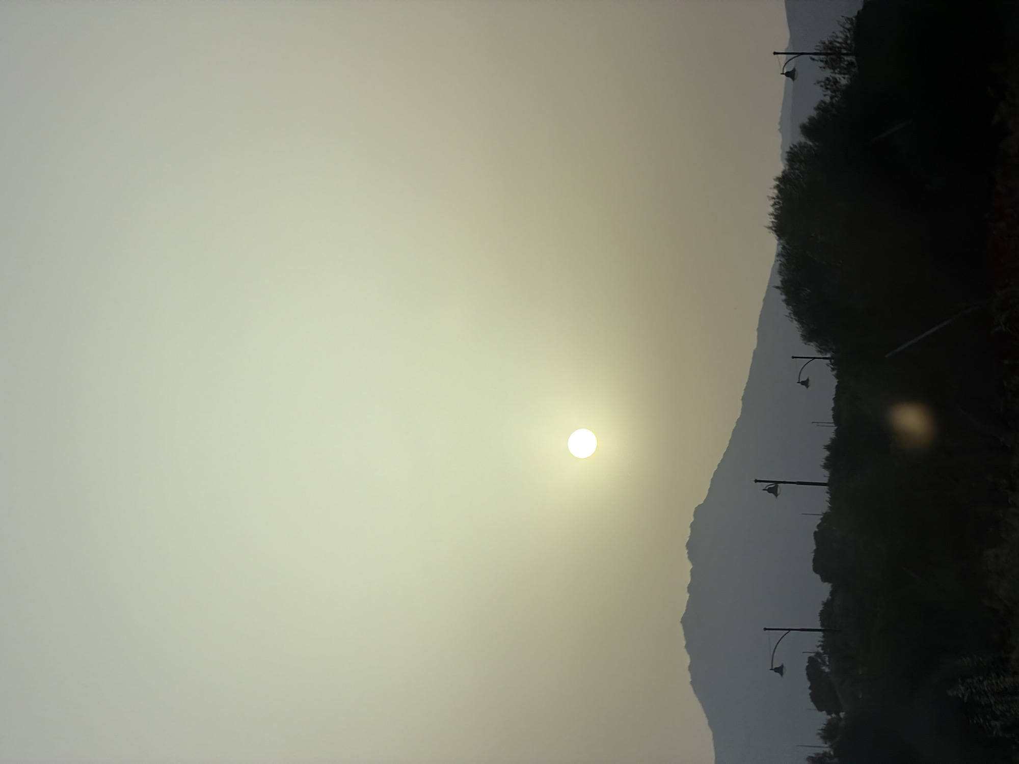 The image depicts a silhouetted landscape under a hazy, pale yellow sky, likely during sunrise or sunset. A bright, diffused sun is visible in the upper center of the frame. In the middle ground, a dark, jagged mountain range forms the horizon. In the foreground, a dark, dense band of vegetation is punctuated by several tall, dark lampposts with decorative tops. The overall impression is one of a rural or semi-rural outskirts, possibly on the periphery of Olbia, Italy. The atmospheric haze obscures finer details of the landscape and vegetation, contributing to a soft, muted visual quality.