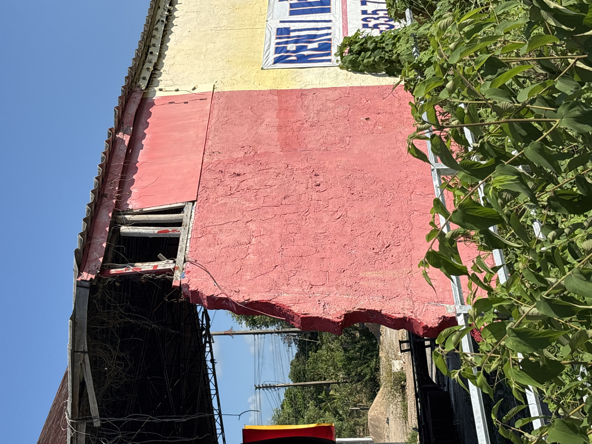An exterior perspective shows a deteriorated, multi-story building with evidence of red, tan, and yellow paint. The building's infrastructure reveals exposed wooden beams, suggesting neglect and potential structural damage. Dense foliage obscures portions of the building and adjacent areas. Outside, urban elements such as utility poles and power lines are visible. The location is Baan Chaweng, Thailand.