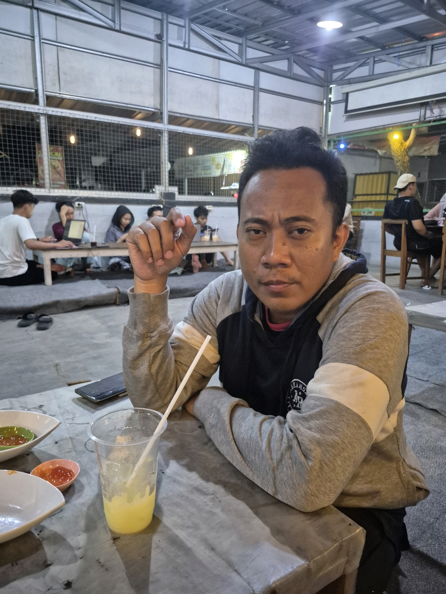 In Pabelan, Indonesia, a man is captured in a medium shot, seated at a table. He is wearing a grey and black hooded sweatshirt, with his arms resting on the table. In front of him, a clear plastic cup contains a yellow beverage with ice and a straw. Two small bowls, one with a green sauce and another with a red sauce, are also on the table. The background reveals an open-air dining area with metal roofing and seating for multiple patrons. Several other individuals are visible in the background, some engaged with laptops and dining. The overall lighting suggests it is evening or nighttime.